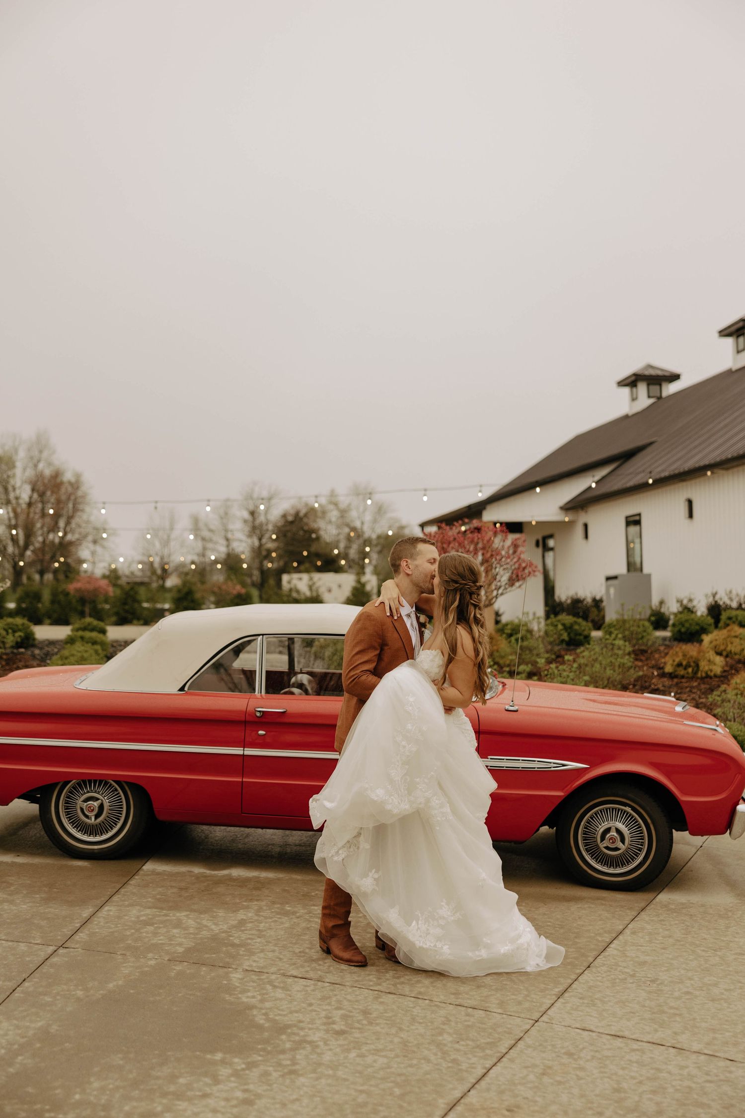 Wedding couple poses with vintage red car outside white building on overcast day.
