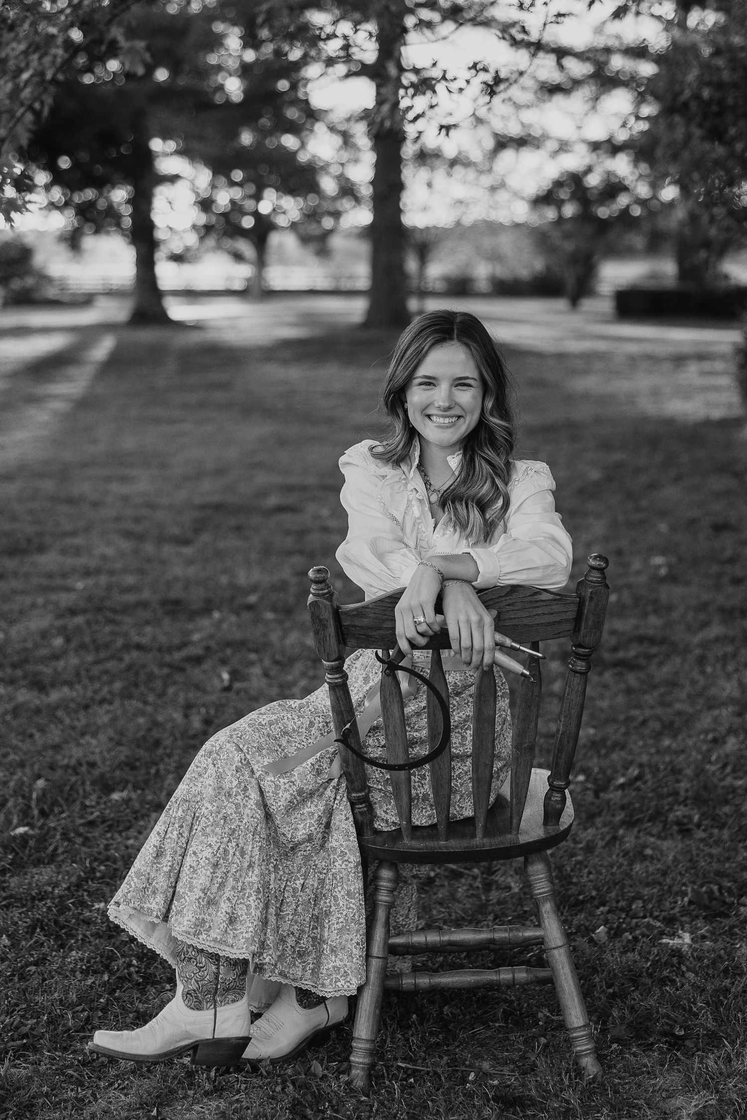 Black and white portrait of someone sitting on a vintage wooden chair in a grassy field.