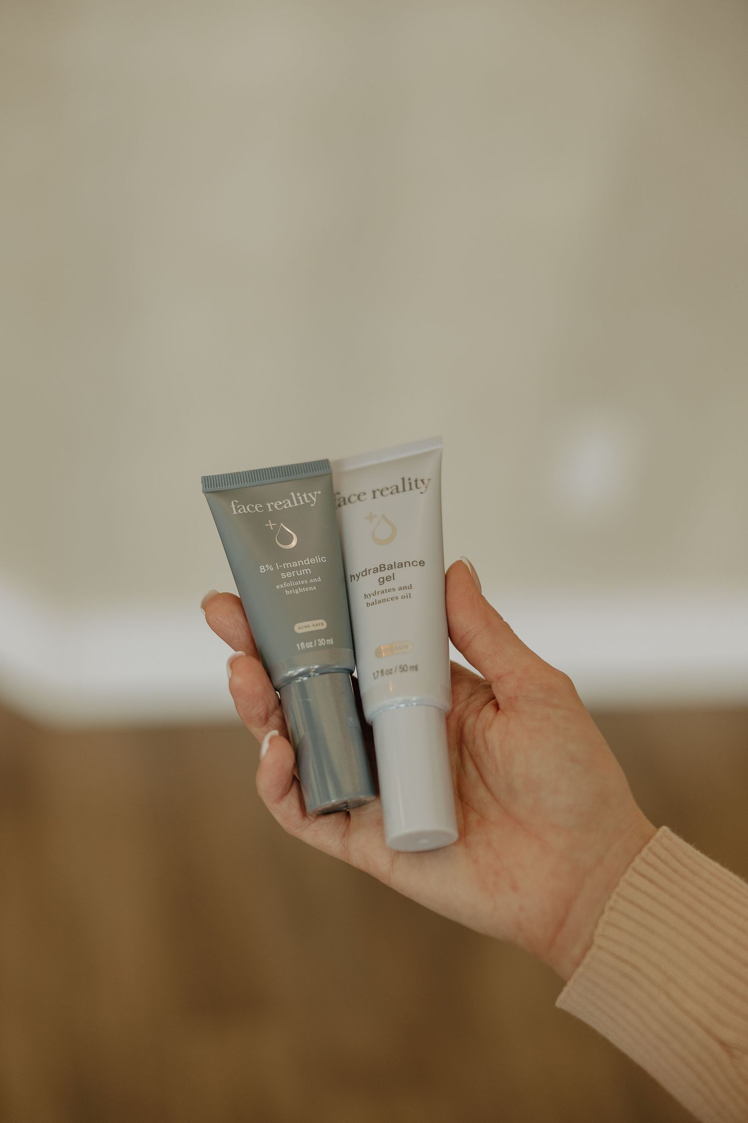 Close up of hand holding two skincare product tubes against neutral background.