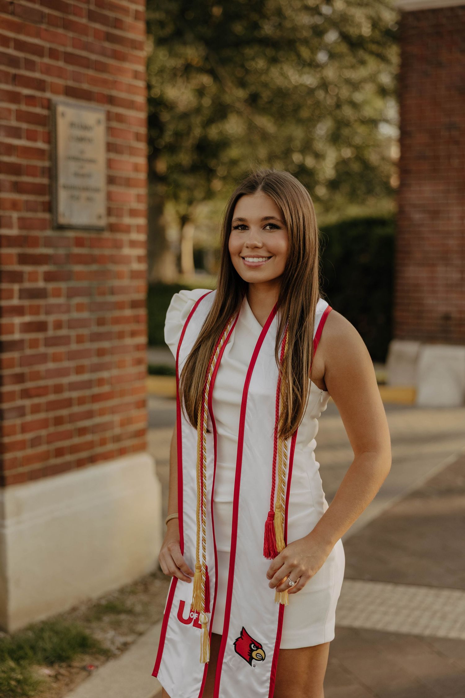 Graduate in white dress with red stoles poses near brick building during golden hour.
