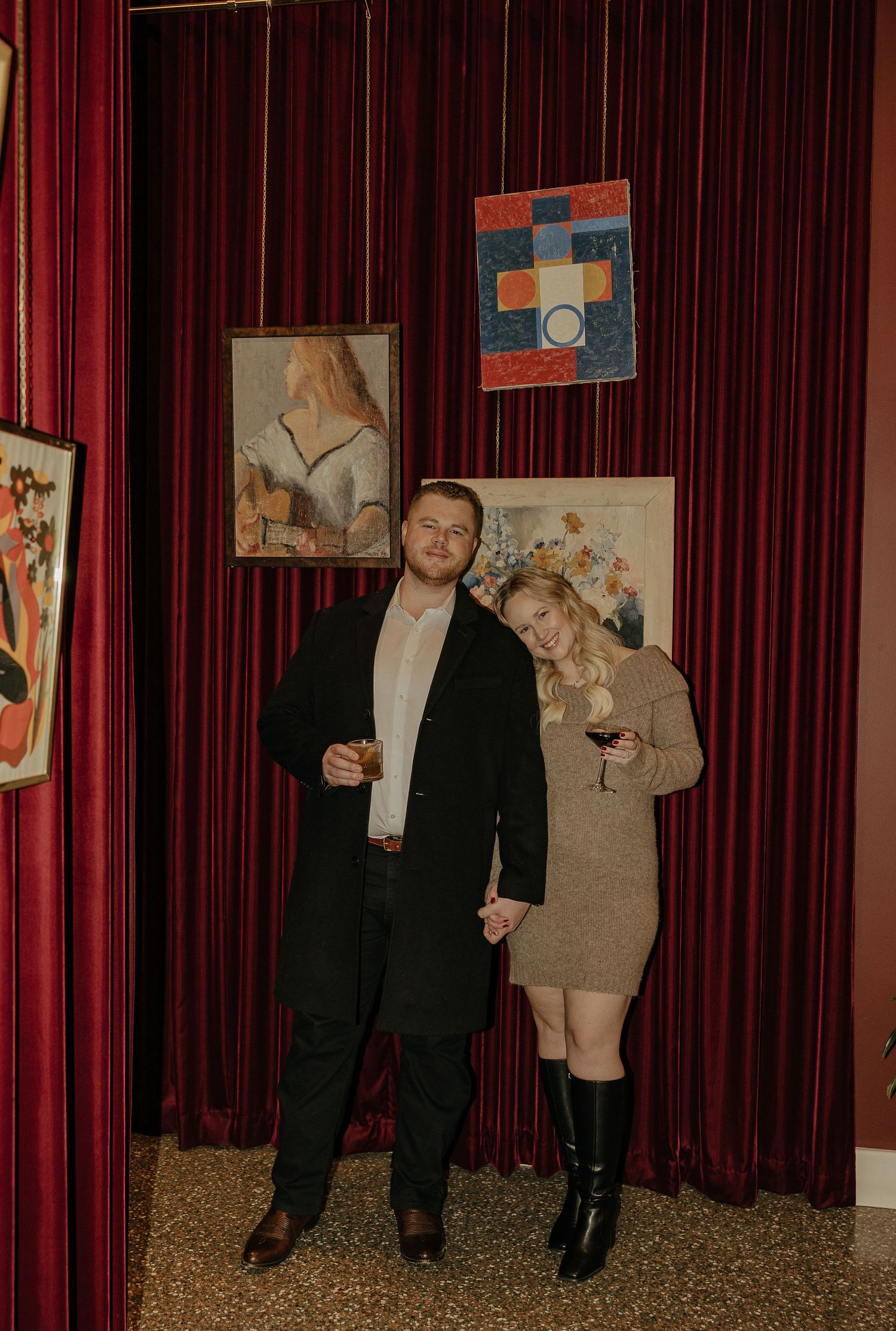 Two people in formal attire stand against red velvet curtains in an elegant room.
