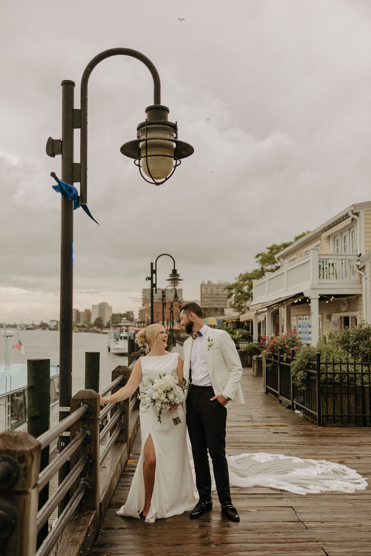 A romantic wedding couple stands on a waterfront boardwalk with vintage lamp posts and city views.