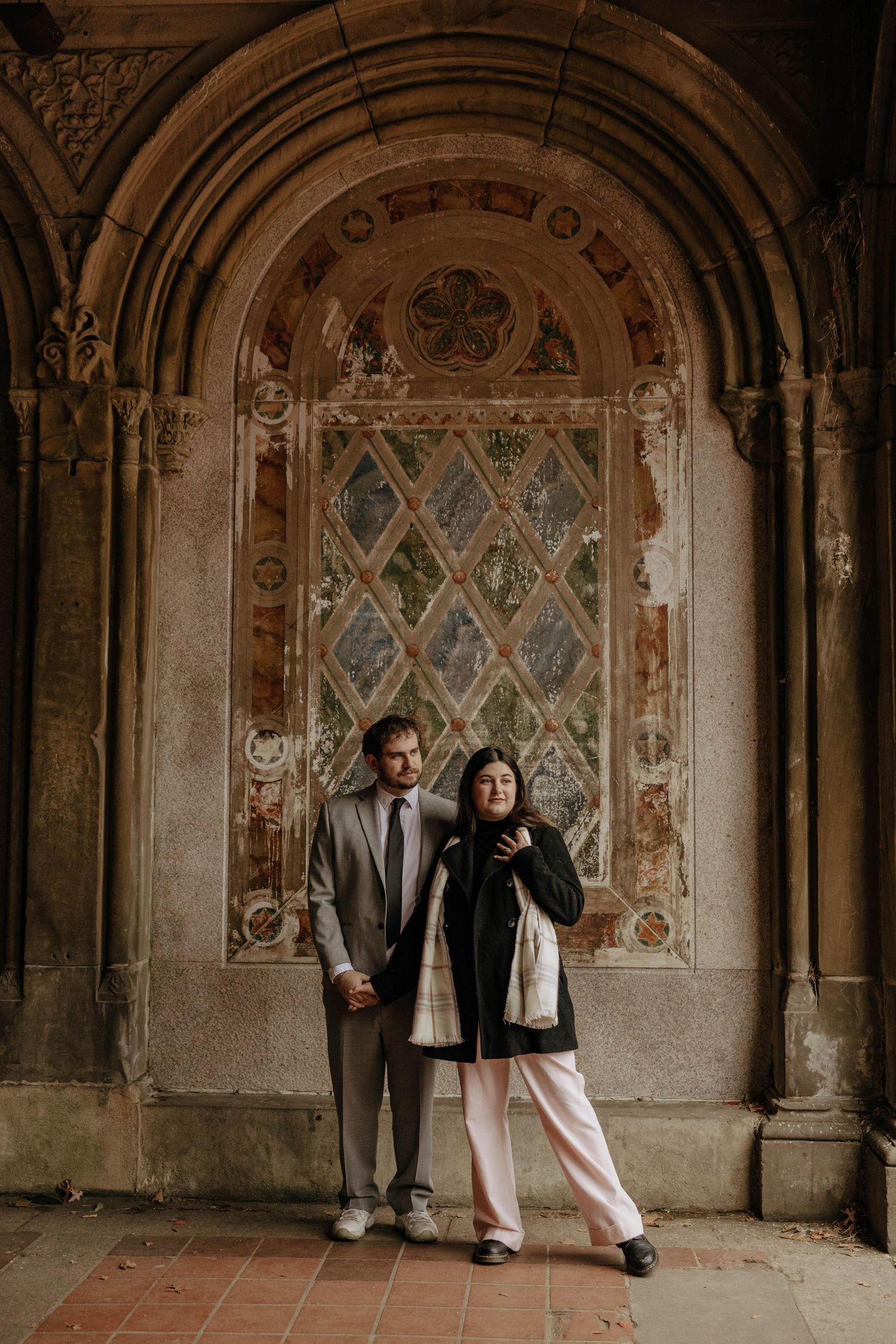 A couple poses in front of an ornate archway with decorative stonework and diamond-patterned detailing at Bethesda Terrace.