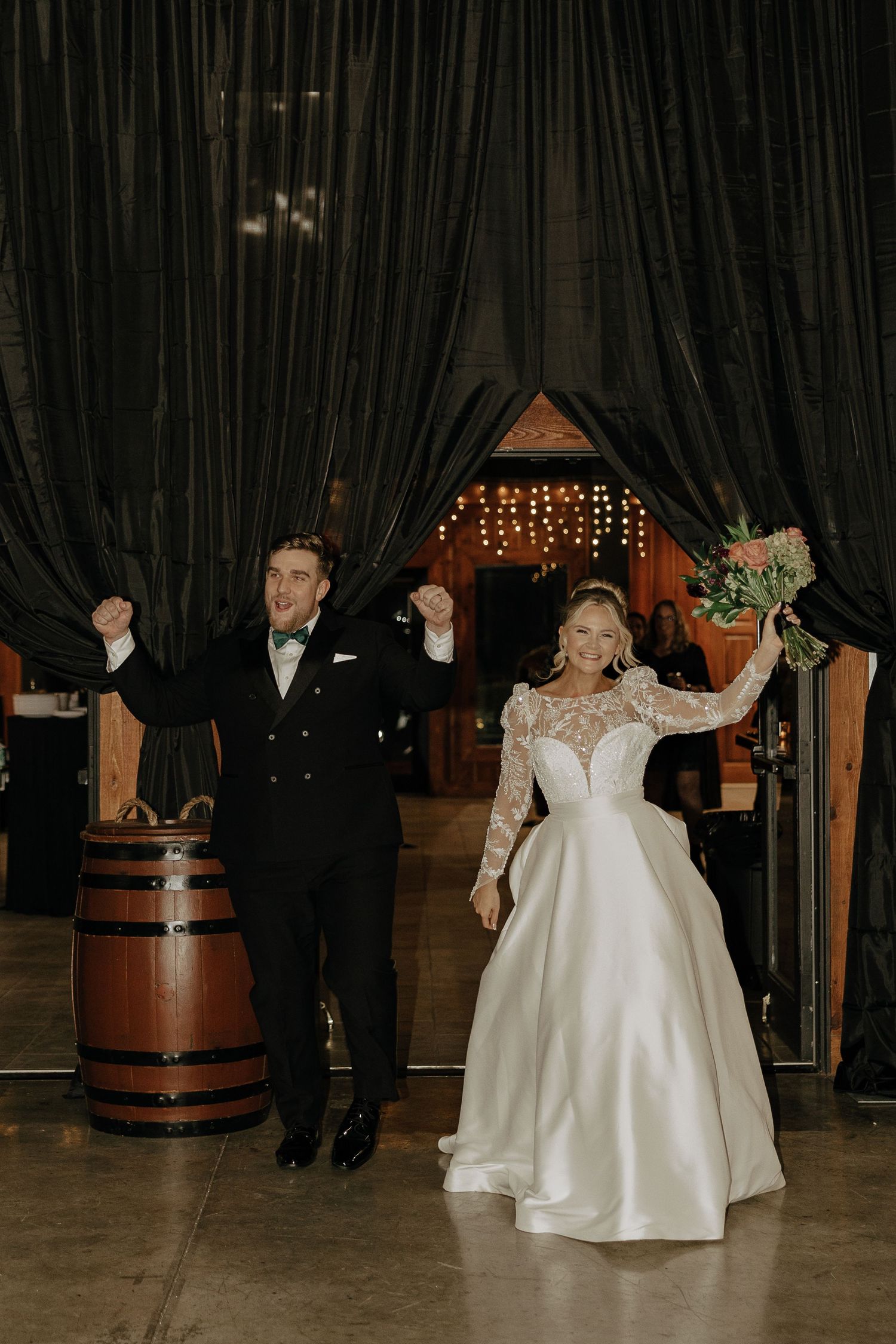 A couple in formal wedding attire dances together at a reception venue decorated with barrels and string lights.