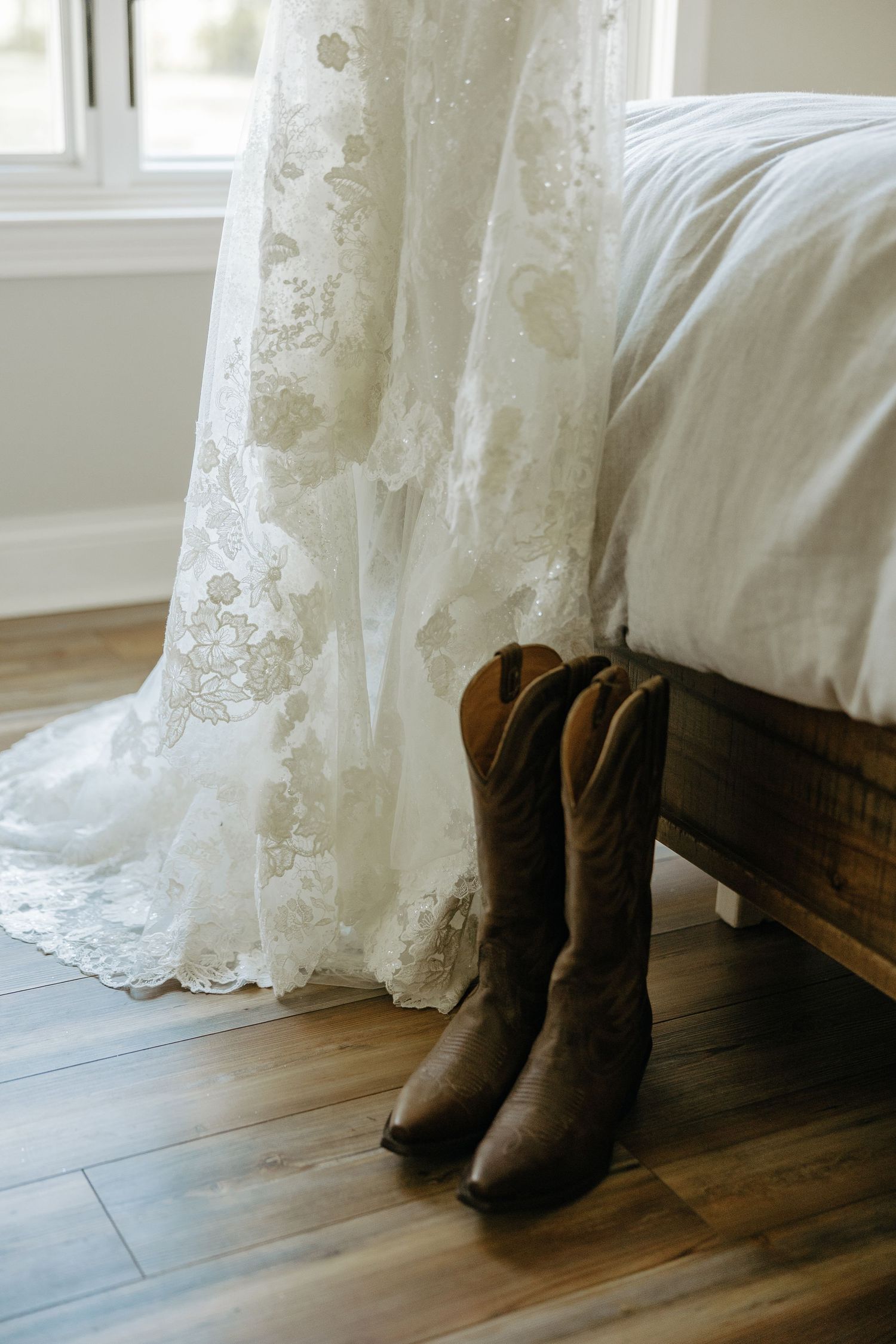 A white lace wedding dress hangs beside brown cowboy boots on a hardwood floor.