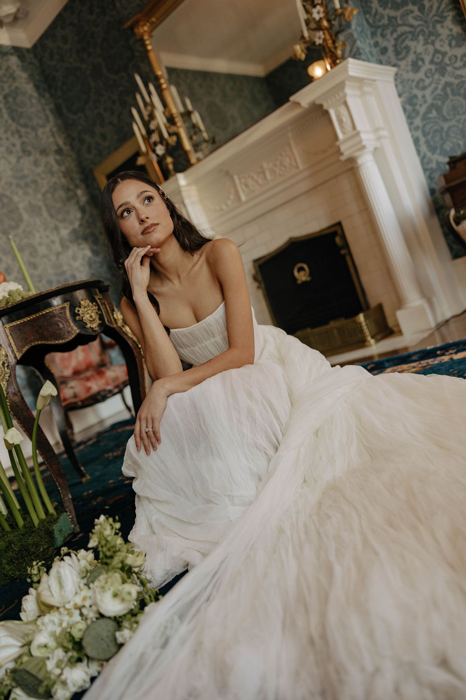 A white wedding gown spreads elegantly across blue carpet near an ornate fireplace.