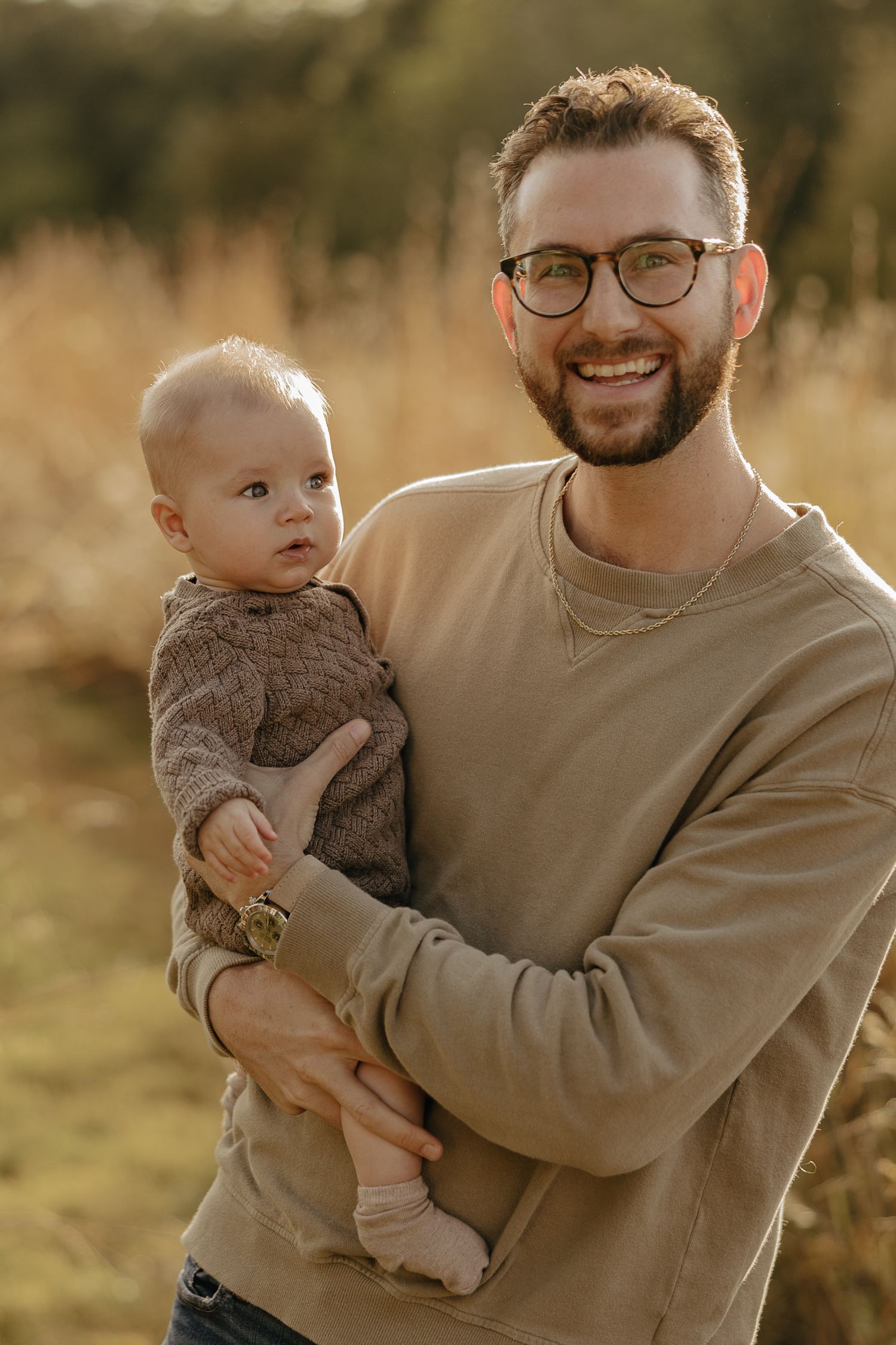 A baby in a brown sweater being held in a golden-lit outdoor setting.