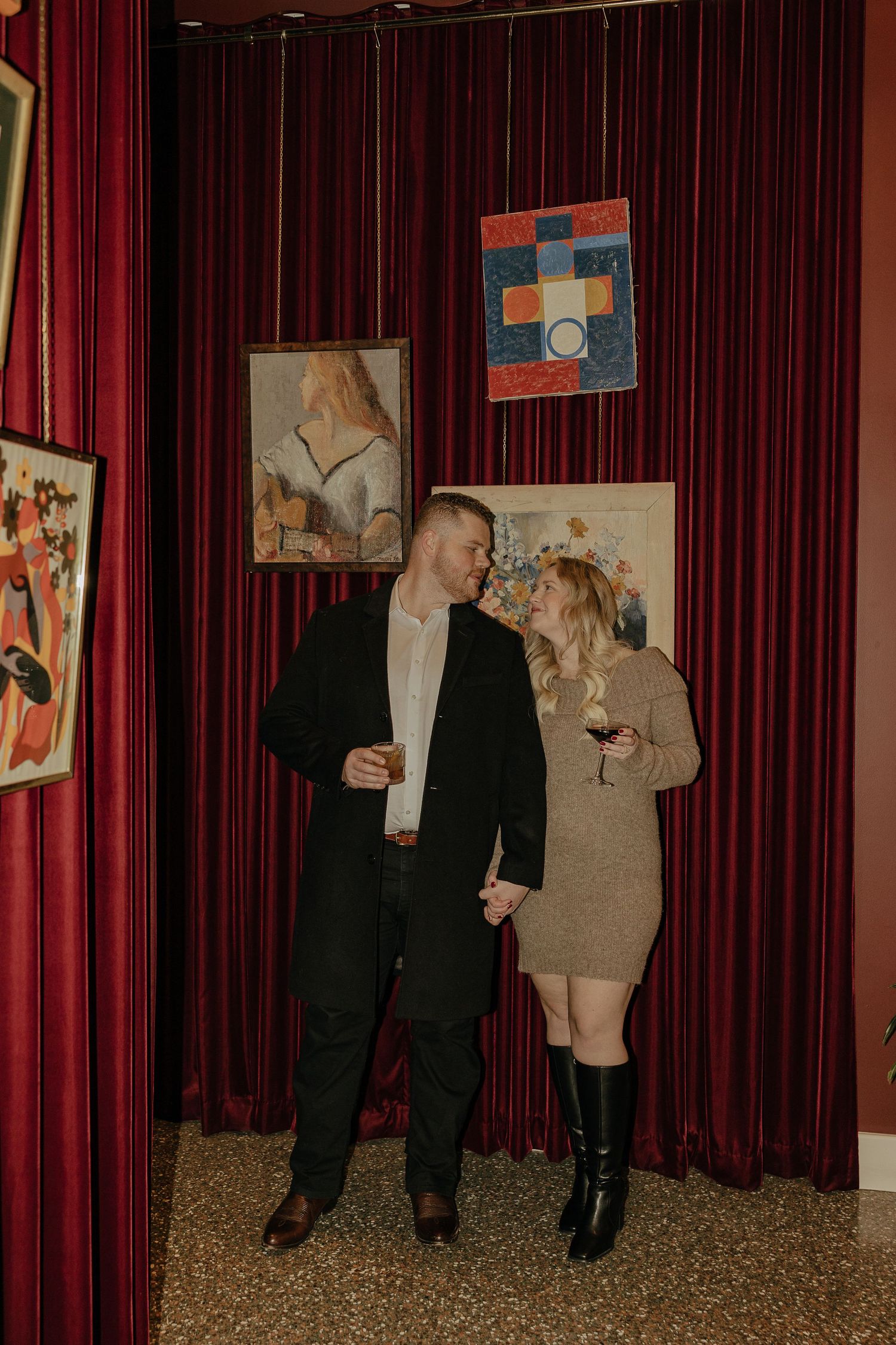 Two people pose against red velvet curtains in an art gallery setting.