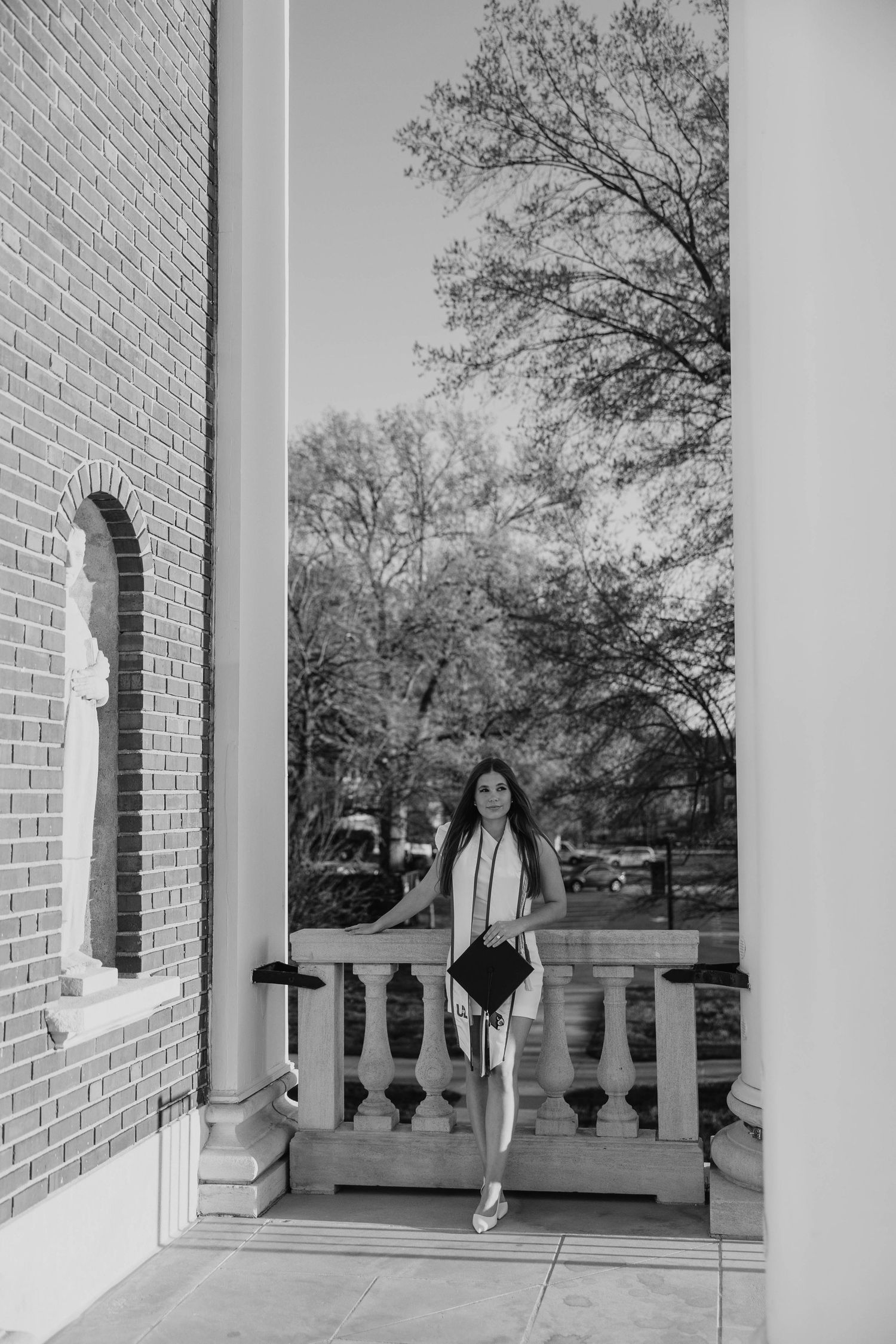 A figure poses on a classical brick porch with white columns and balustrades in a black and white photo.