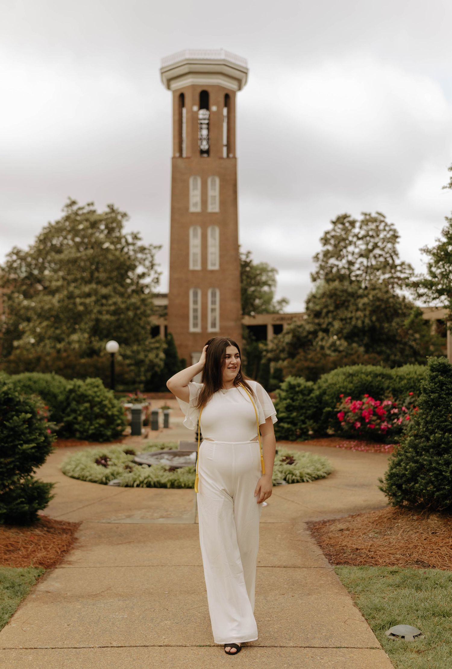 White graduation dress photographed in front of brick bell tower and landscaped grounds.