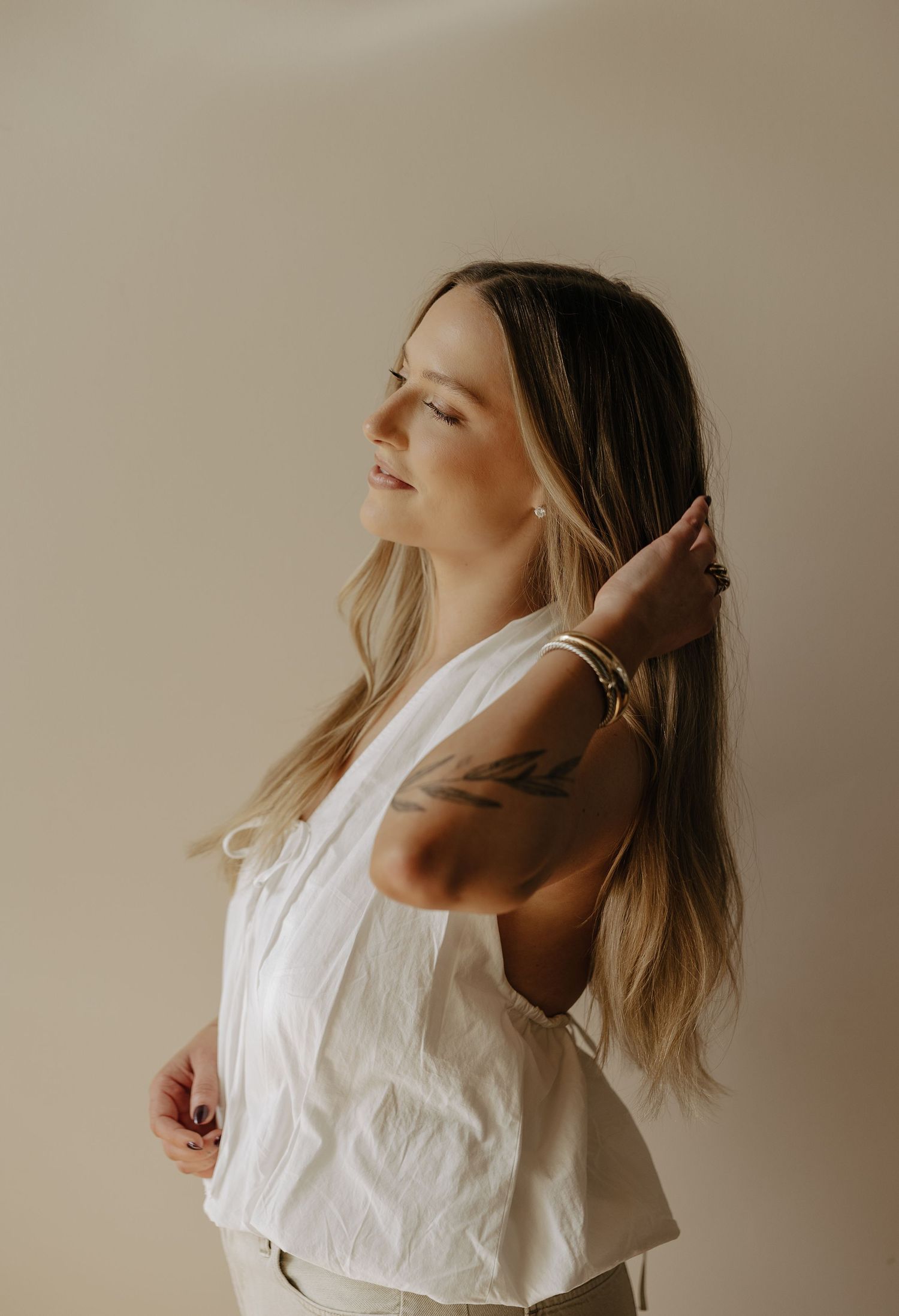 Young woman in white top with arm tattoo poses with hand in long blonde hair against neutral background.
