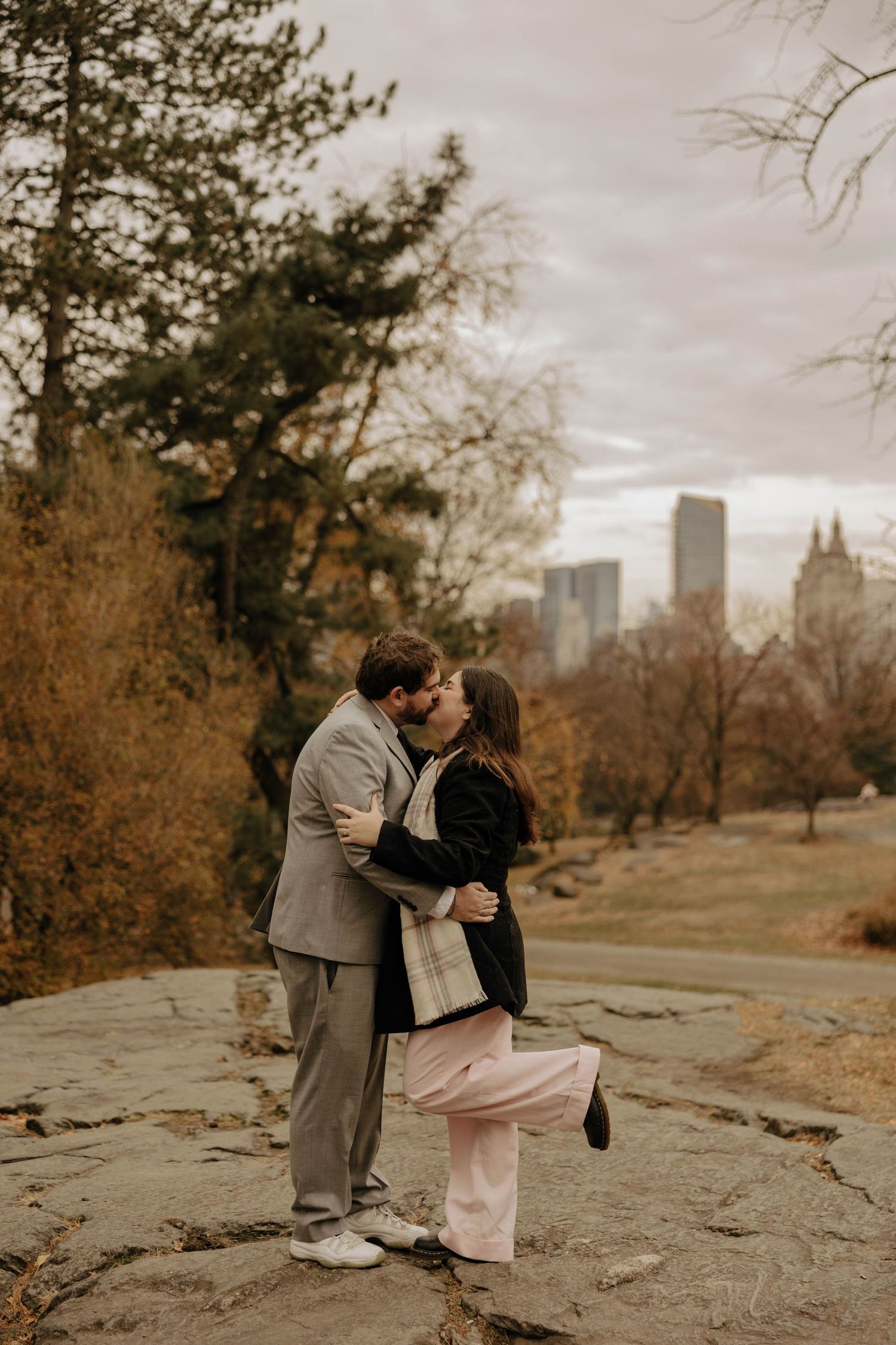 A couple shares a romantic moment in a park with the city skyline visible in the autumn background.