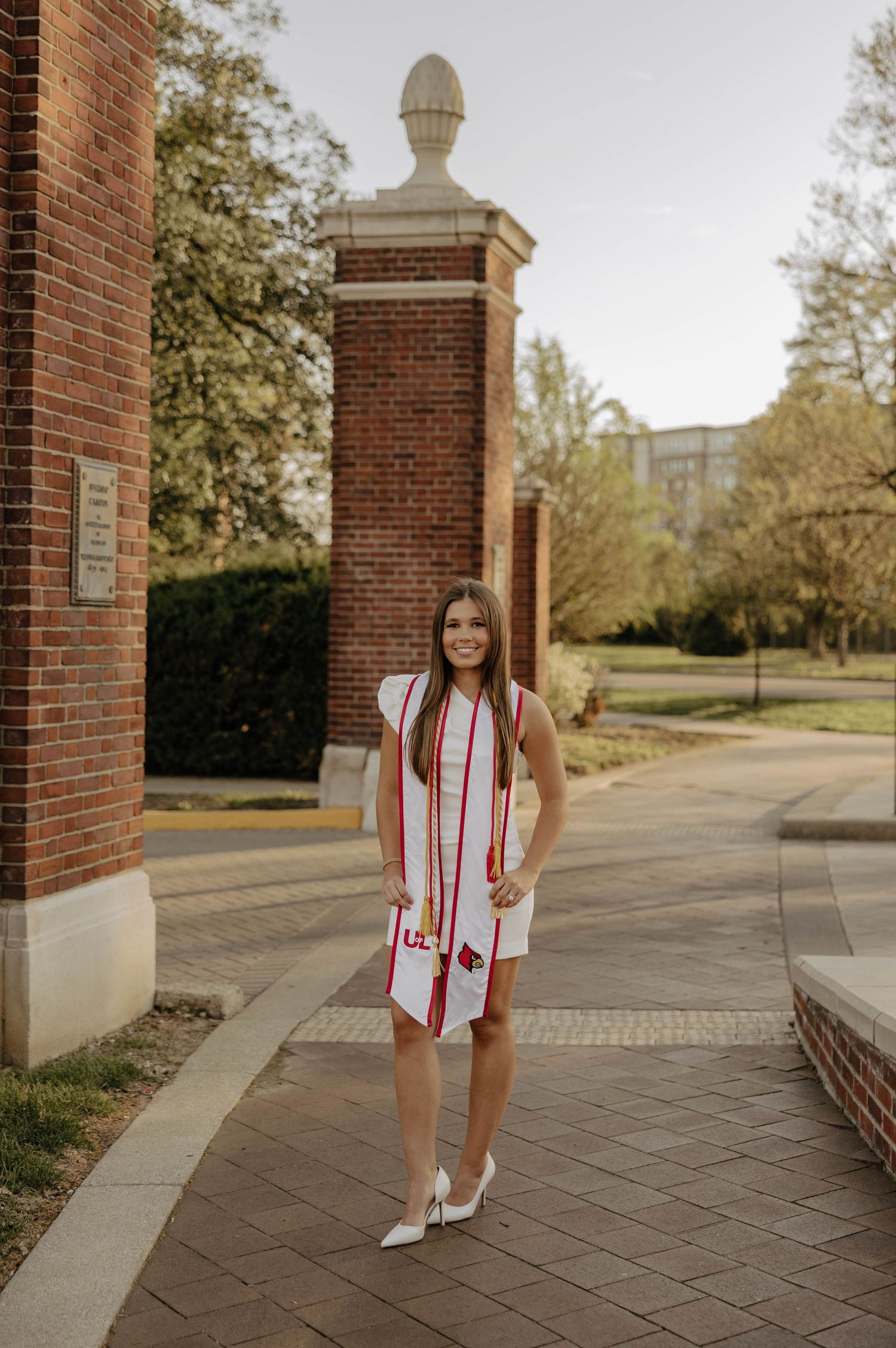 Graduate in white dress and red graduation stole walking near brick building.