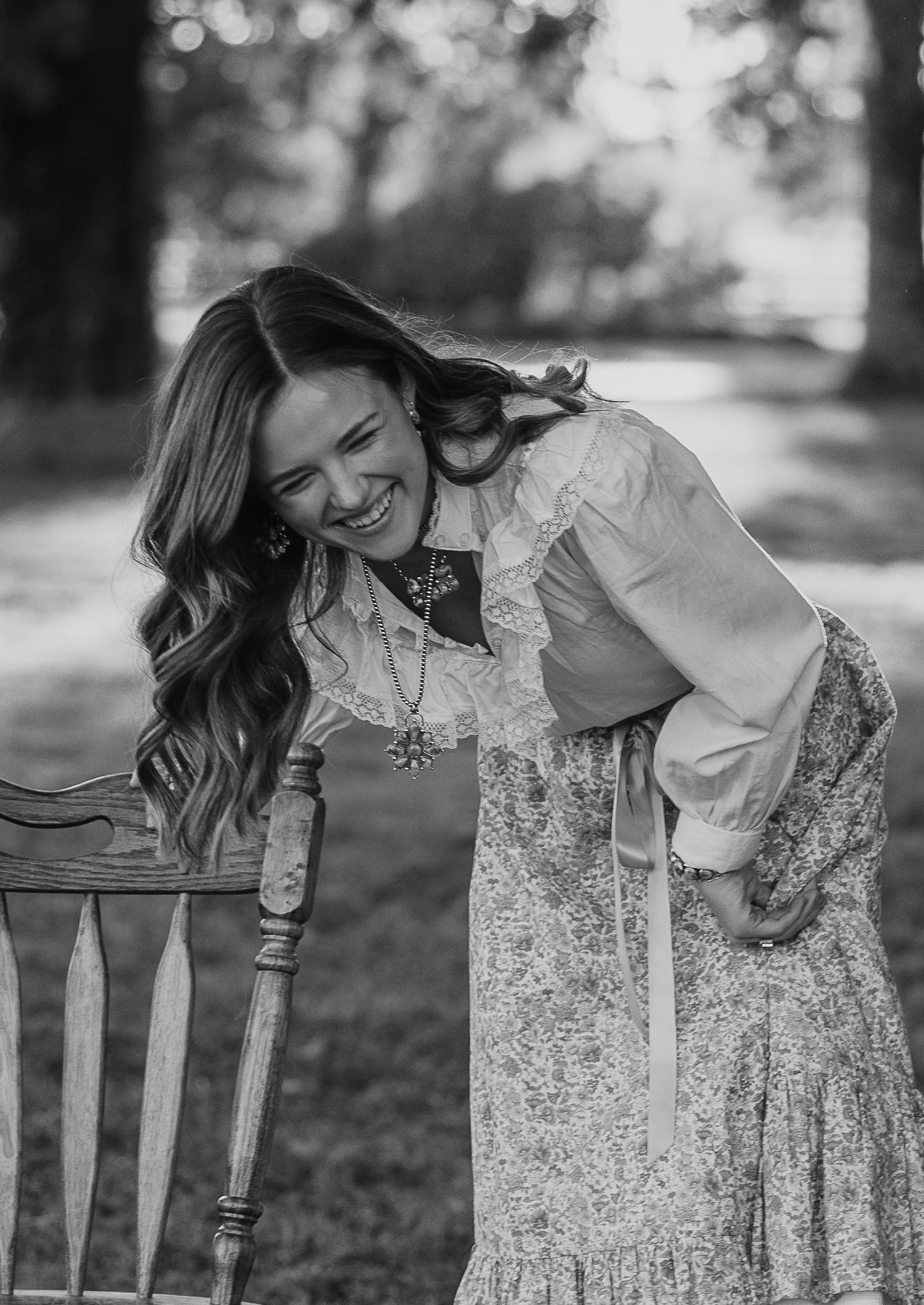 Black and white portrait of someone seated on a wooden chair in a dreamy outdoor setting.
