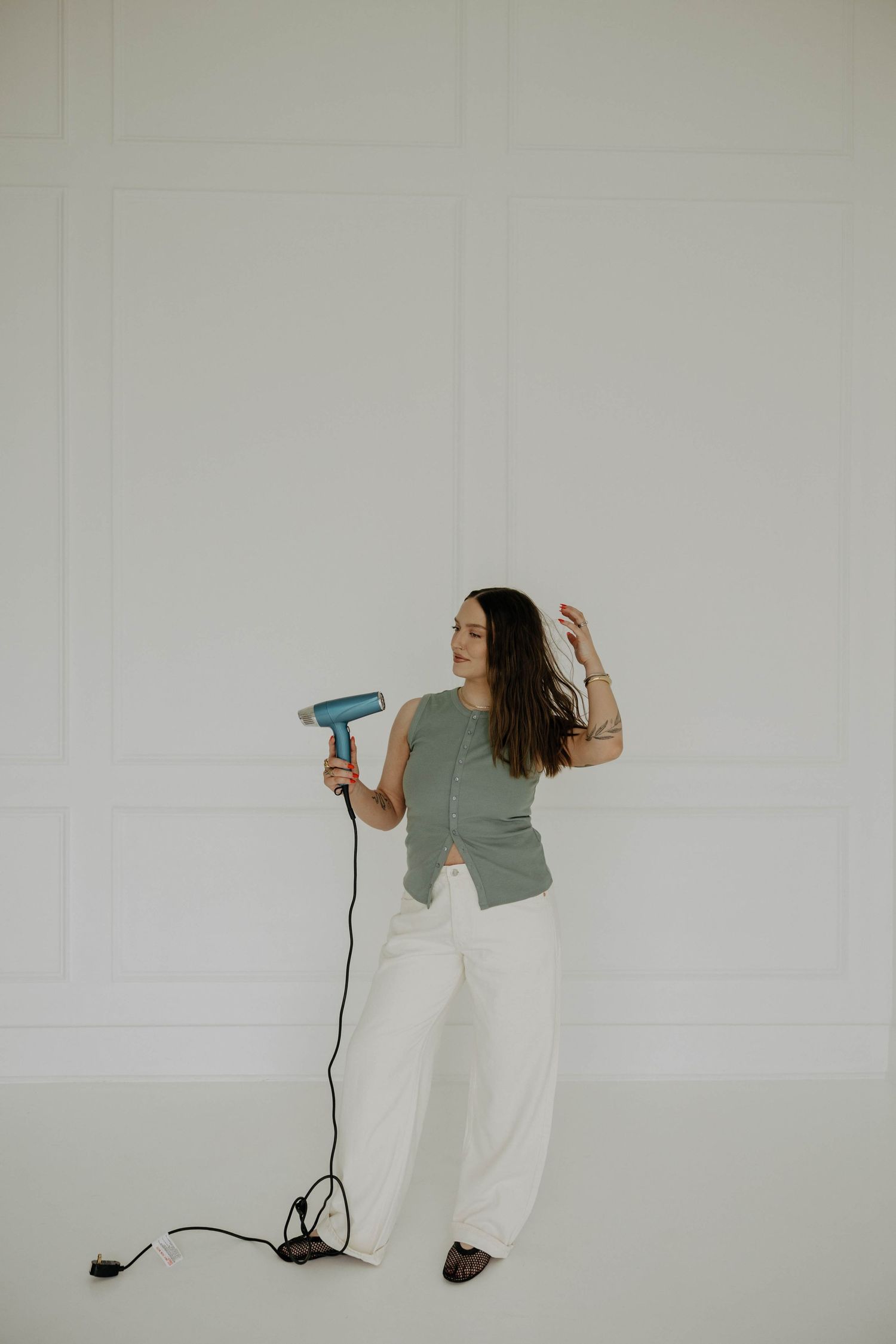 A person in casual attire holds a blue hair dryer while posing against a white wall.