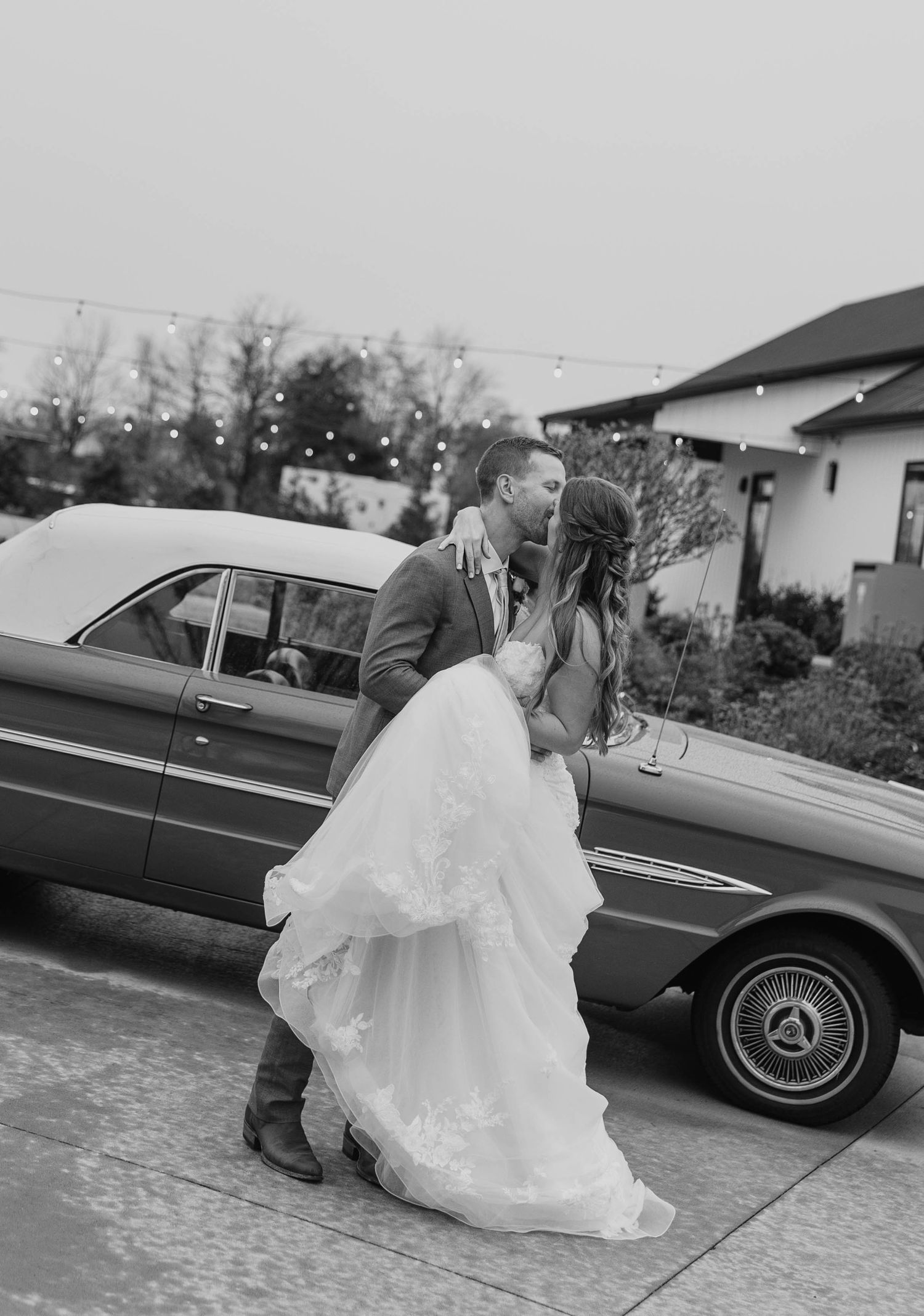 Black and white photo of couple posing by vintage car on their wedding day.