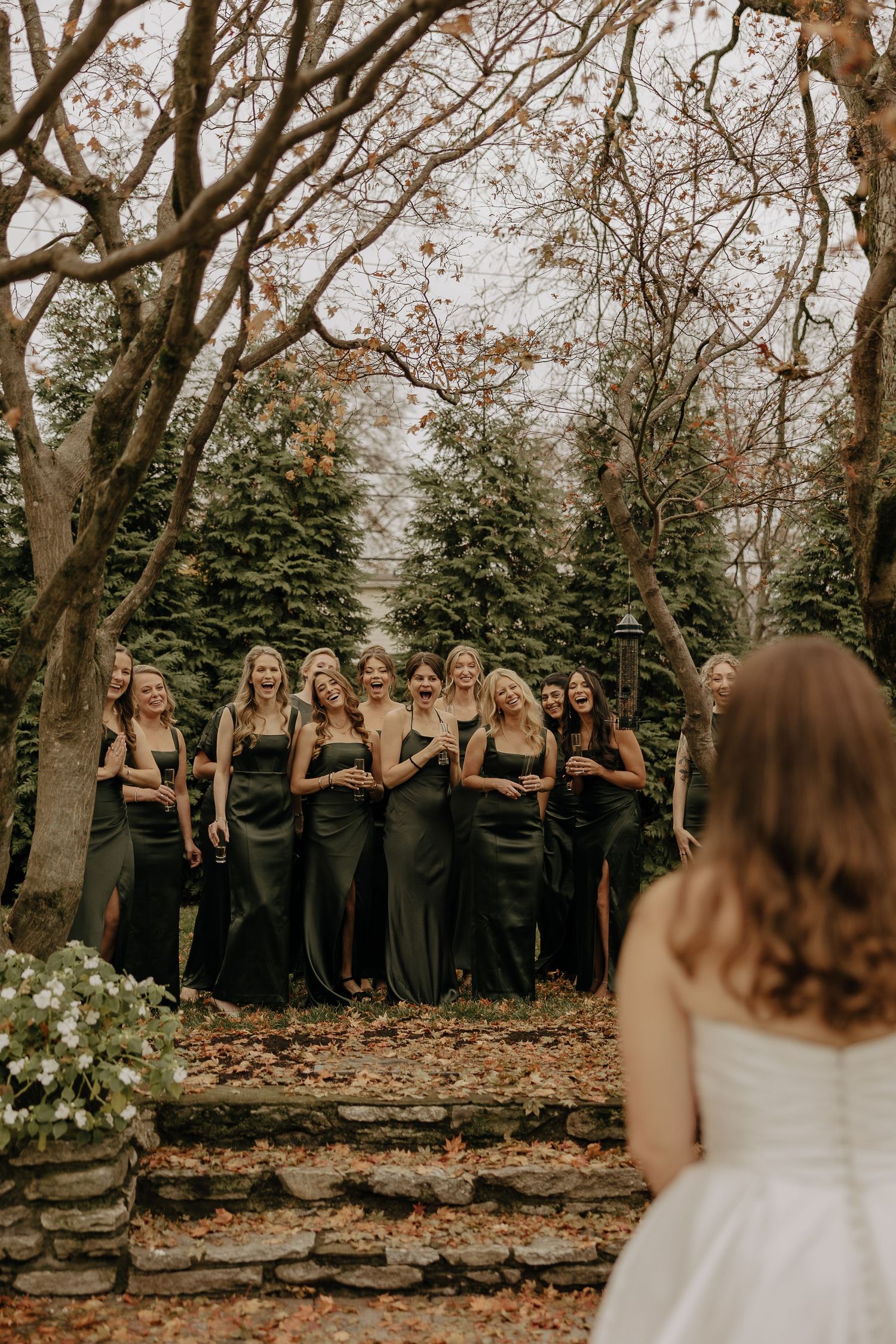 Bridesmaids in black dresses stand together among autumn trees during outdoor wedding ceremony.
