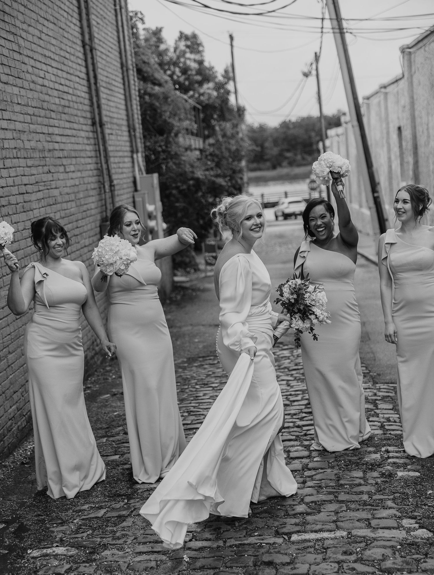 Bridesmaids in light colored dresses pose playfully on cobblestone street.