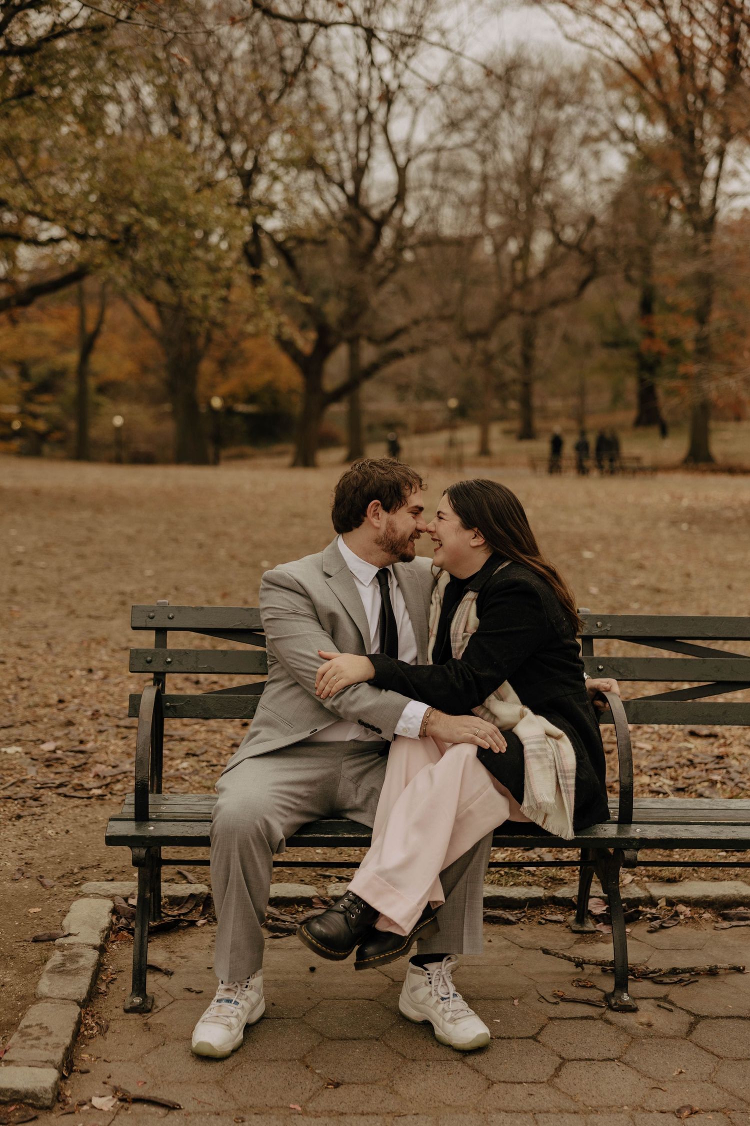 A romantic couple sits close together on a park bench surrounded by autumn trees and fallen leaves.