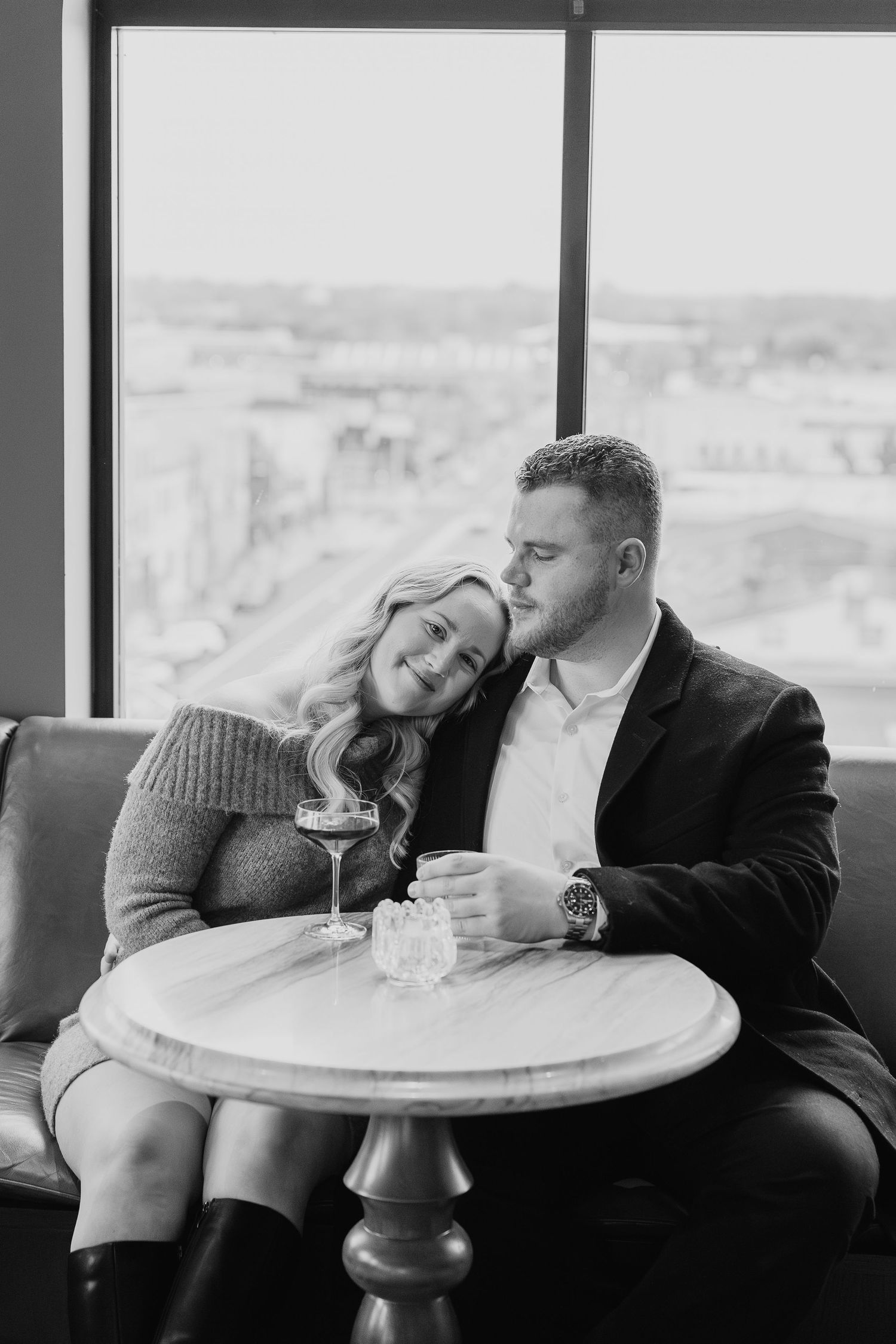 A black and white photo of a couple sharing drinks at a round table by large windows.
