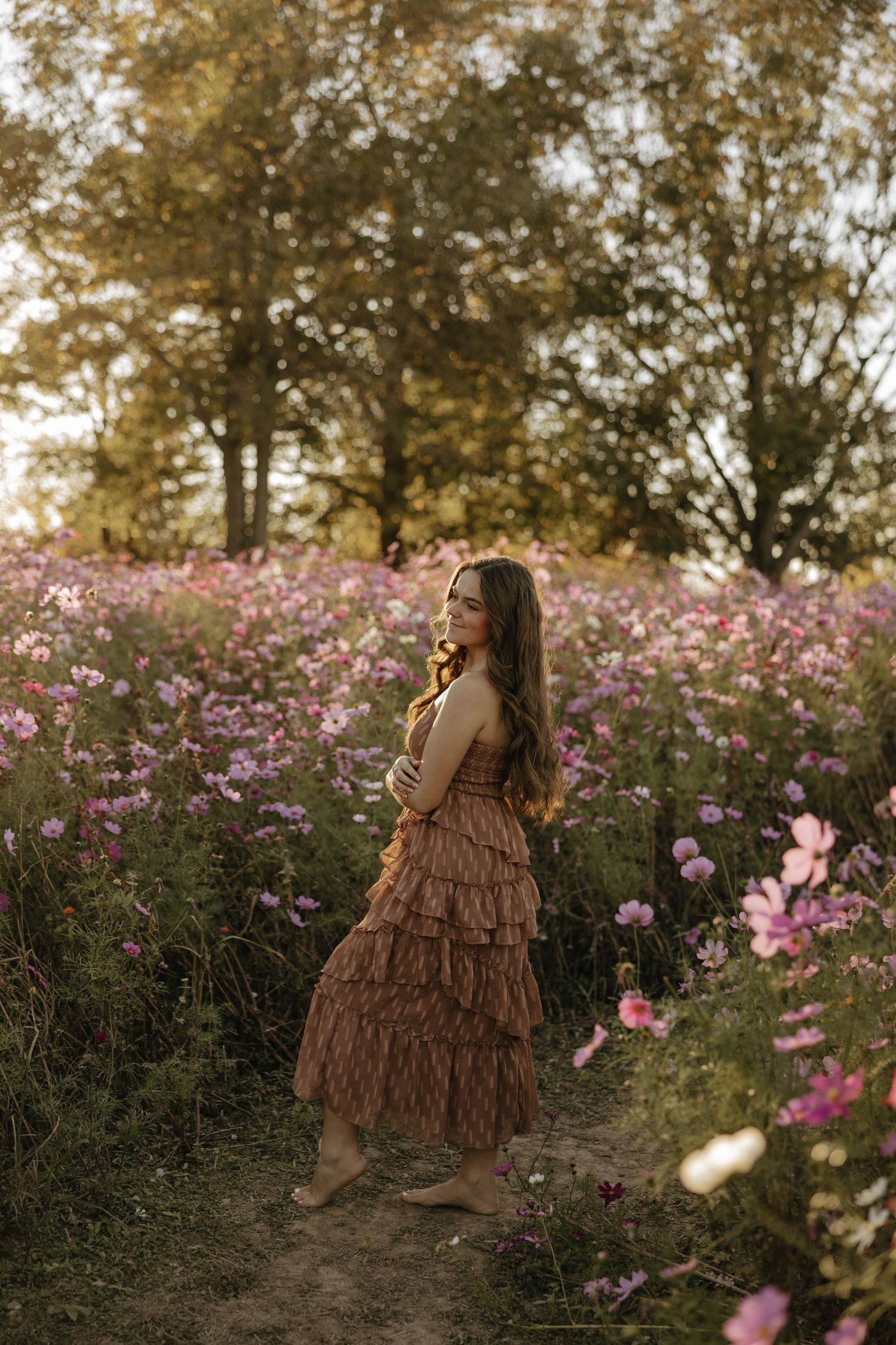 A figure in a tiered brown dress poses among pink wildflowers at sunset.