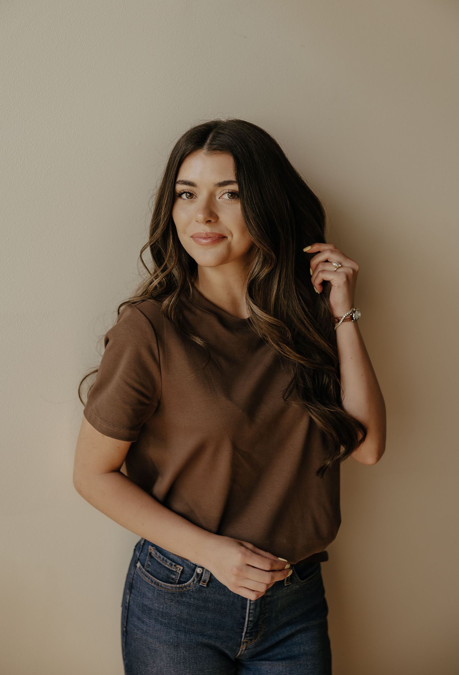 Young woman with long dark wavy hair wearing a brown t-shirt and jeans posing against a beige wall.