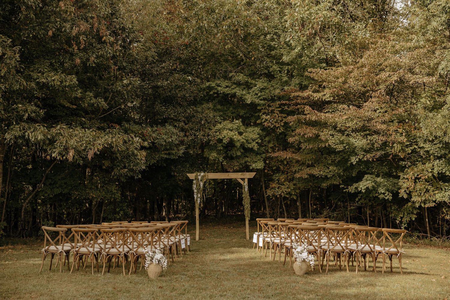 Wooden cross-back chairs set up for an outdoor wedding ceremony in a wooded area.
