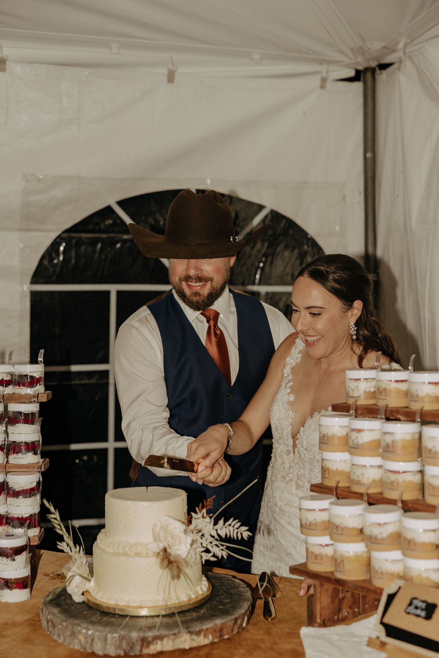 A wedding couple cuts their cake next to a display of desserts under a white tent.