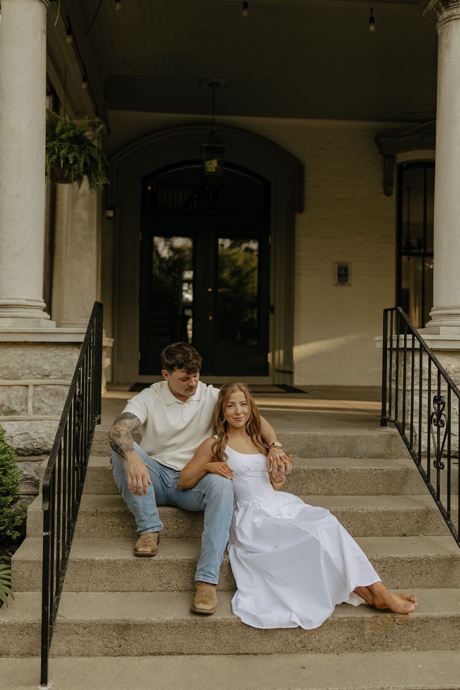 Couple sitting on stone steps of elegant home, woman in white dress, man in jeans and white tee.