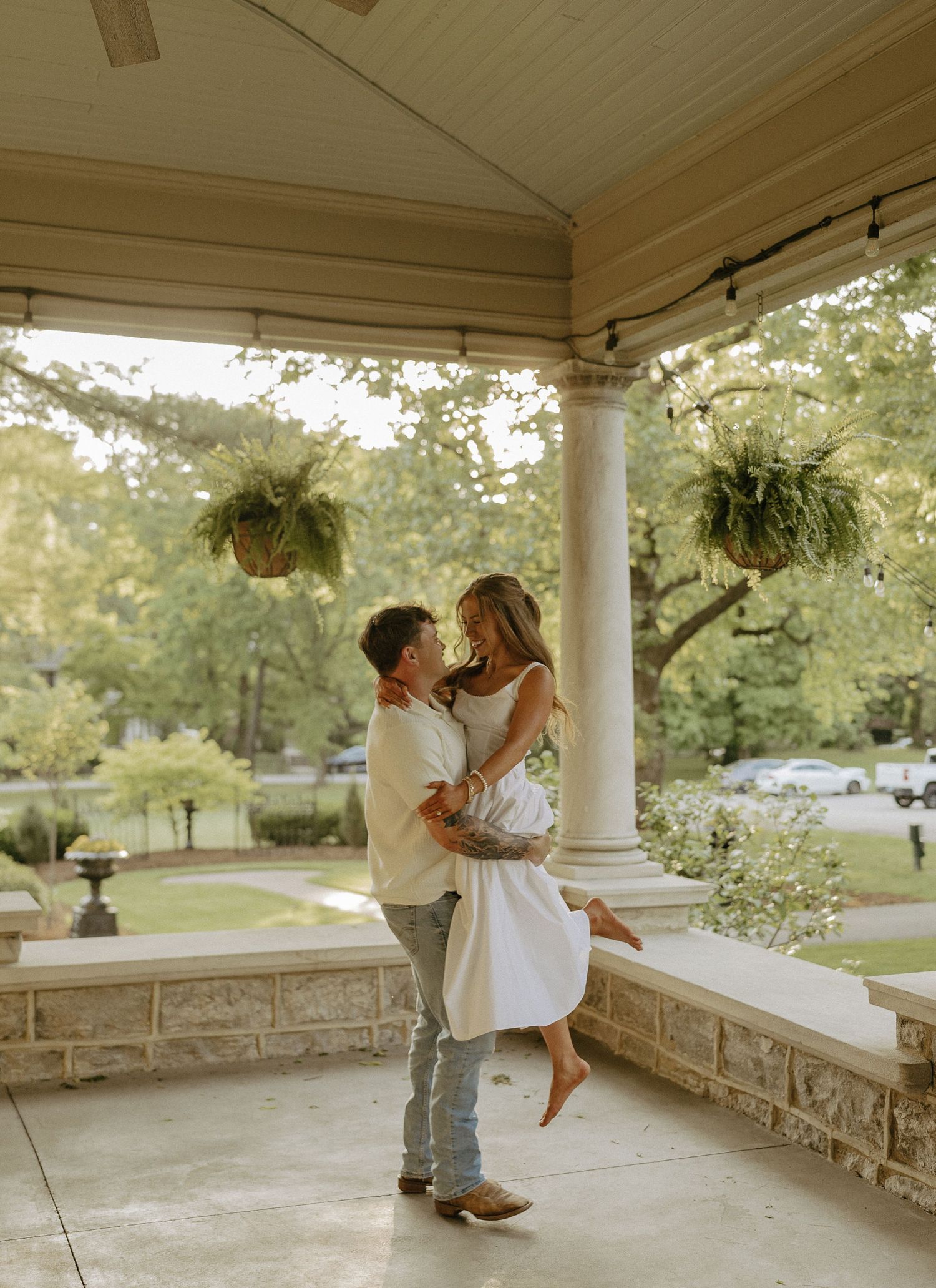 Romantic couple embraces on a elegant covered porch with hanging ferns and white columns.