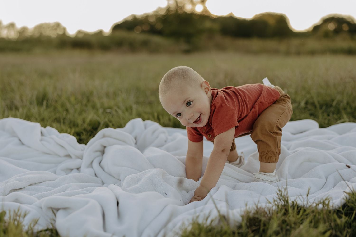Baby crawls on white blanket laid out in green grass field.
