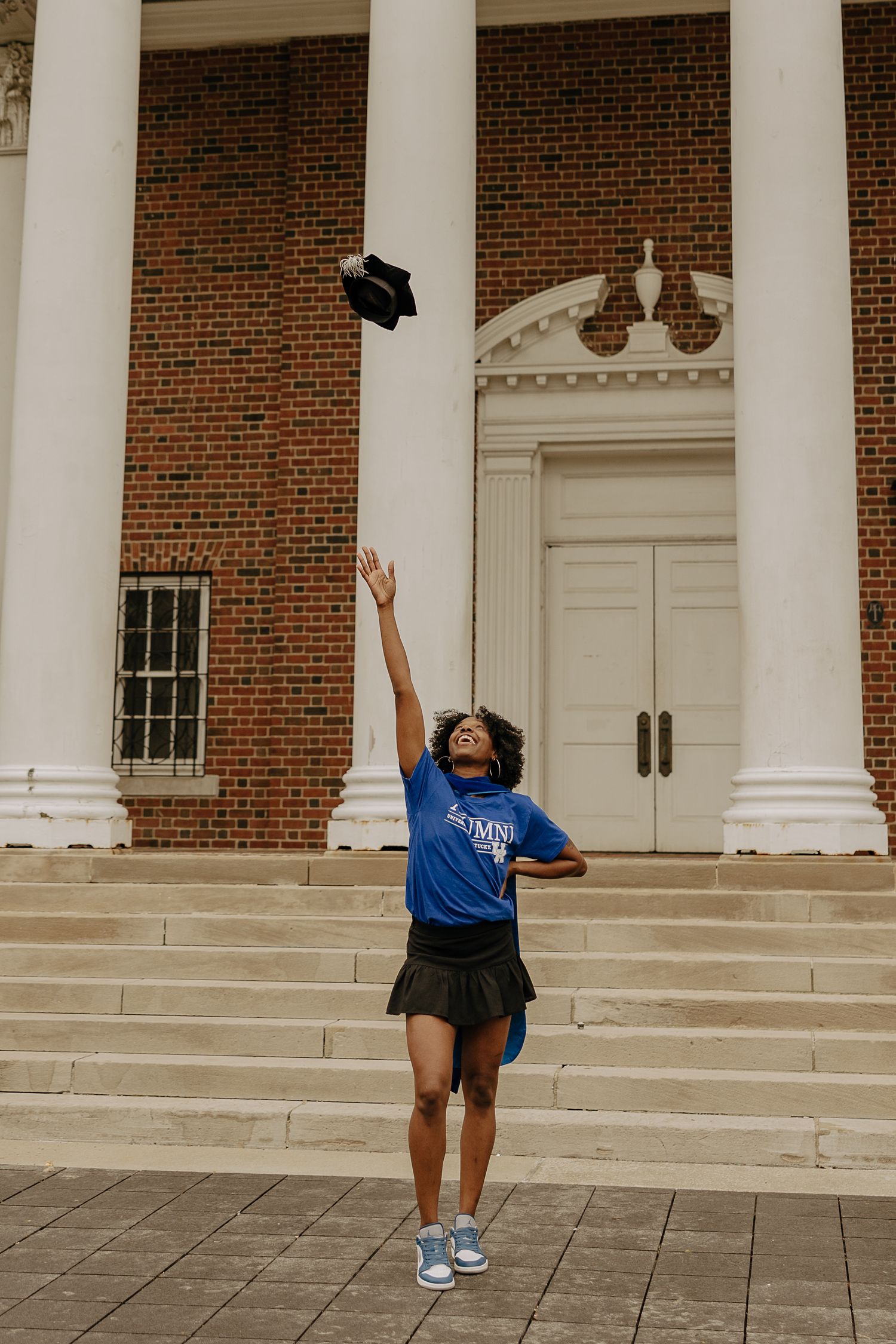 Graduate celebrates on steps of columned brick building throwing cap in celebration.