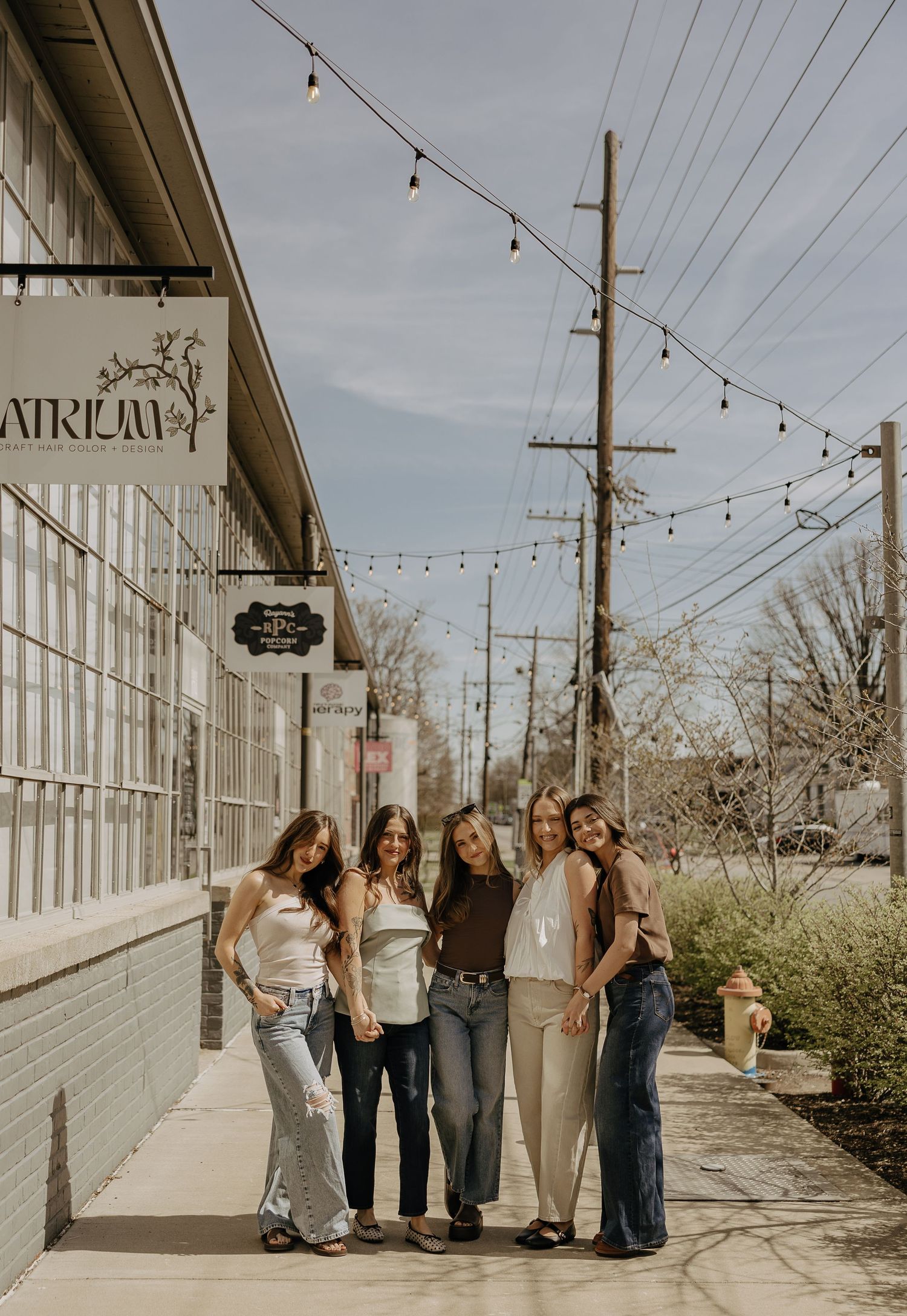 Five women pose together on a sunny sidewalk outside the Atrium venue with string lights overhead.