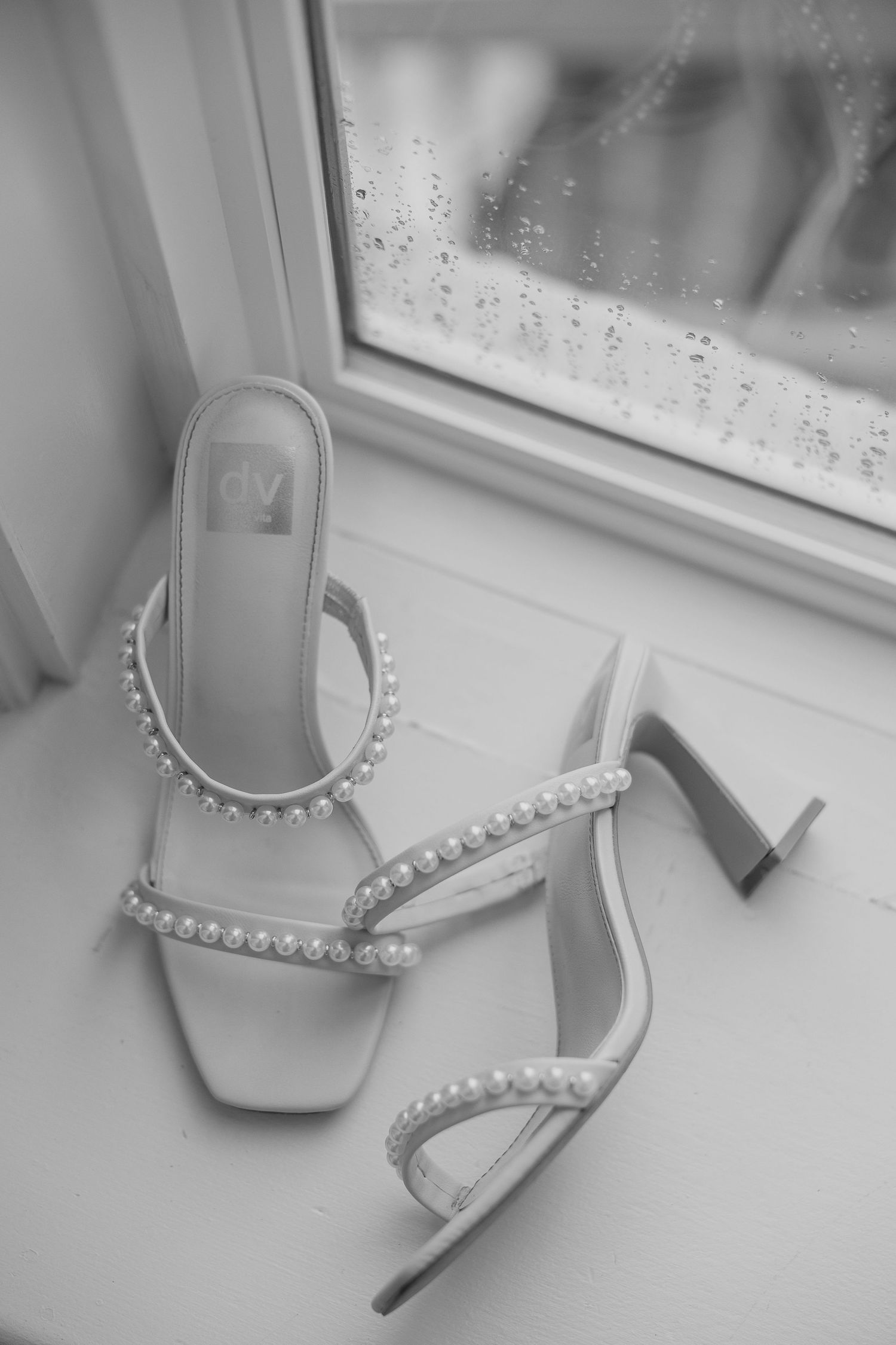 Elegant white pearl-embellished bridal heels displayed near a window with raindrops.