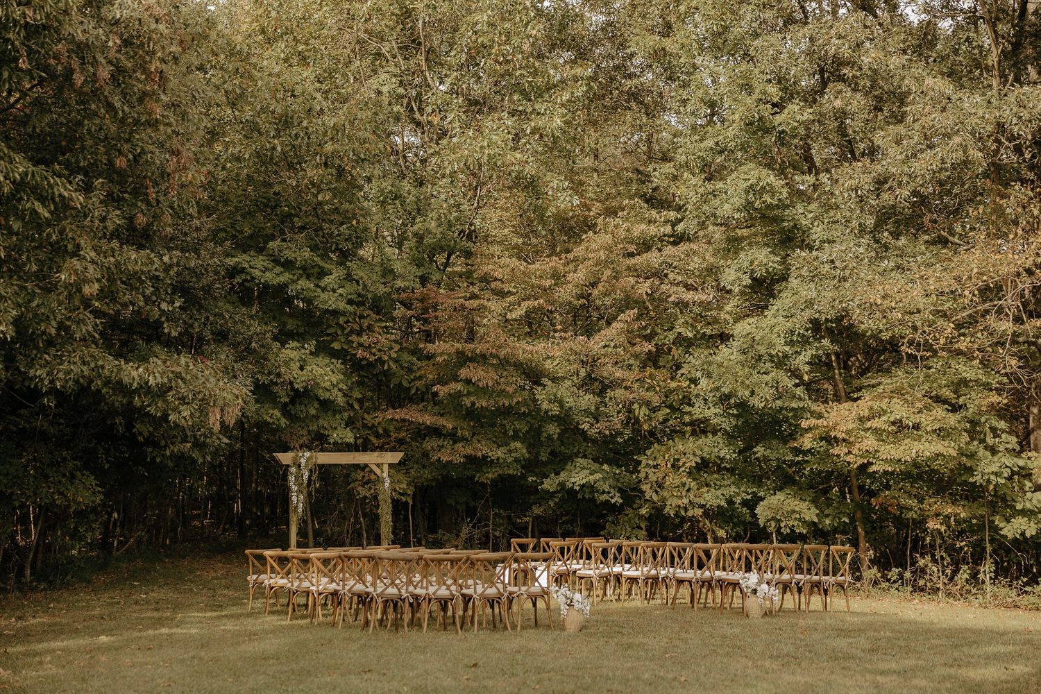 Outdoor wedding ceremony setup with wooden chairs arranged in rows among trees.