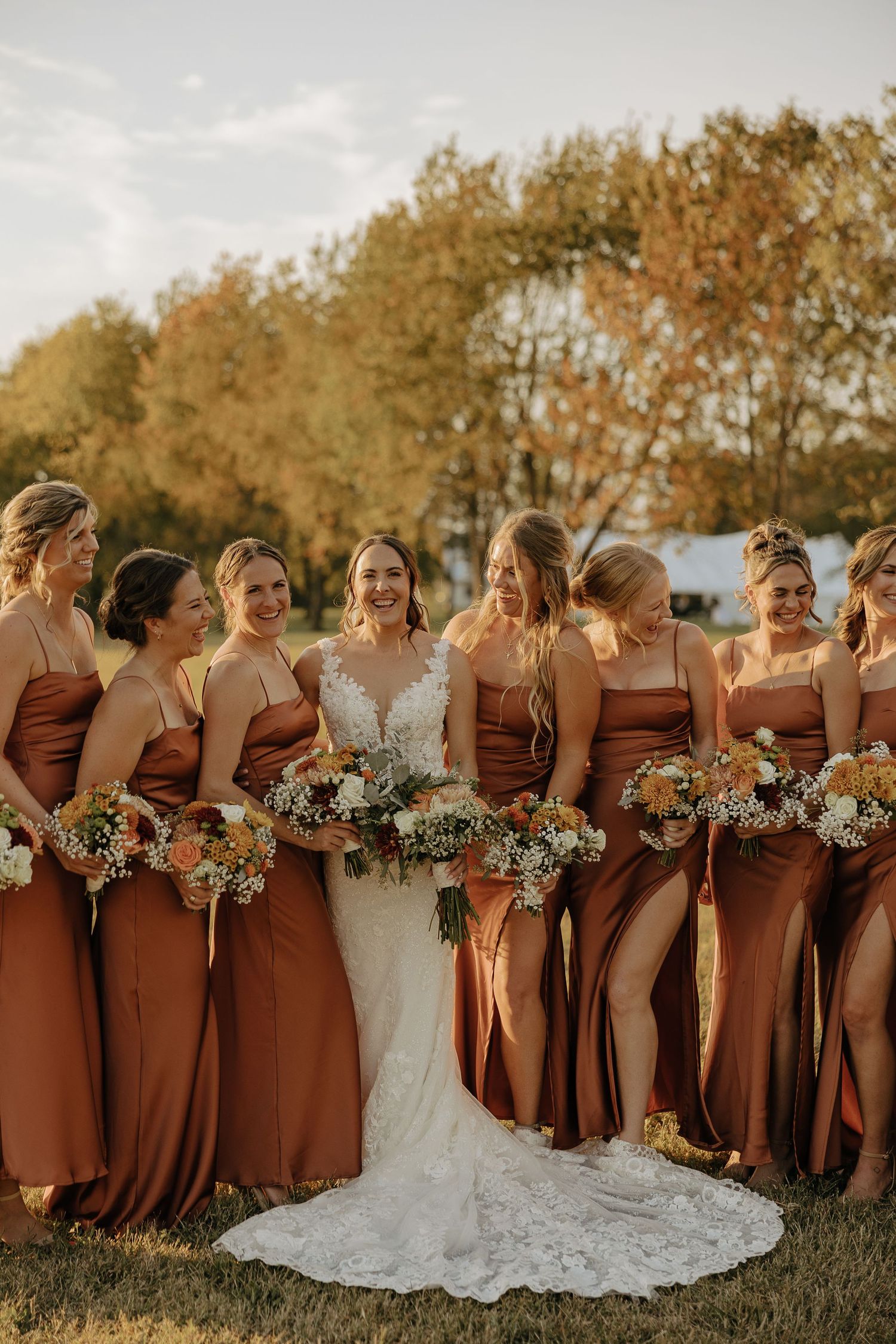 Bridesmaids in matching brown dresses pose together holding floral bouquets in a grassy field.