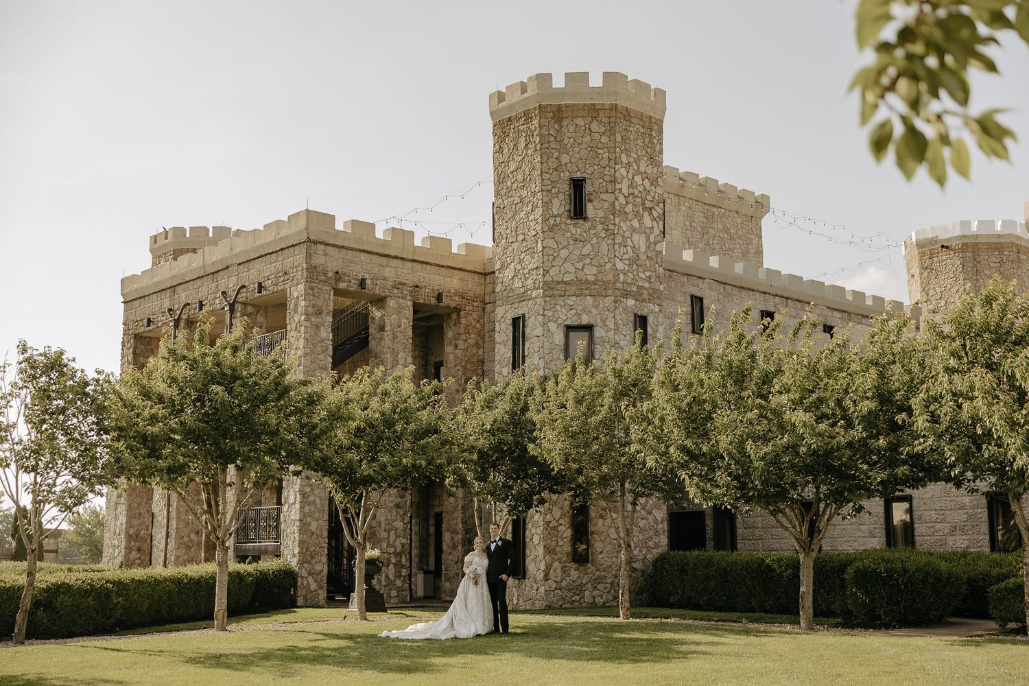 A panoramic view of a historic stone castle with multiple towers and battlements surrounded by green landscaping.