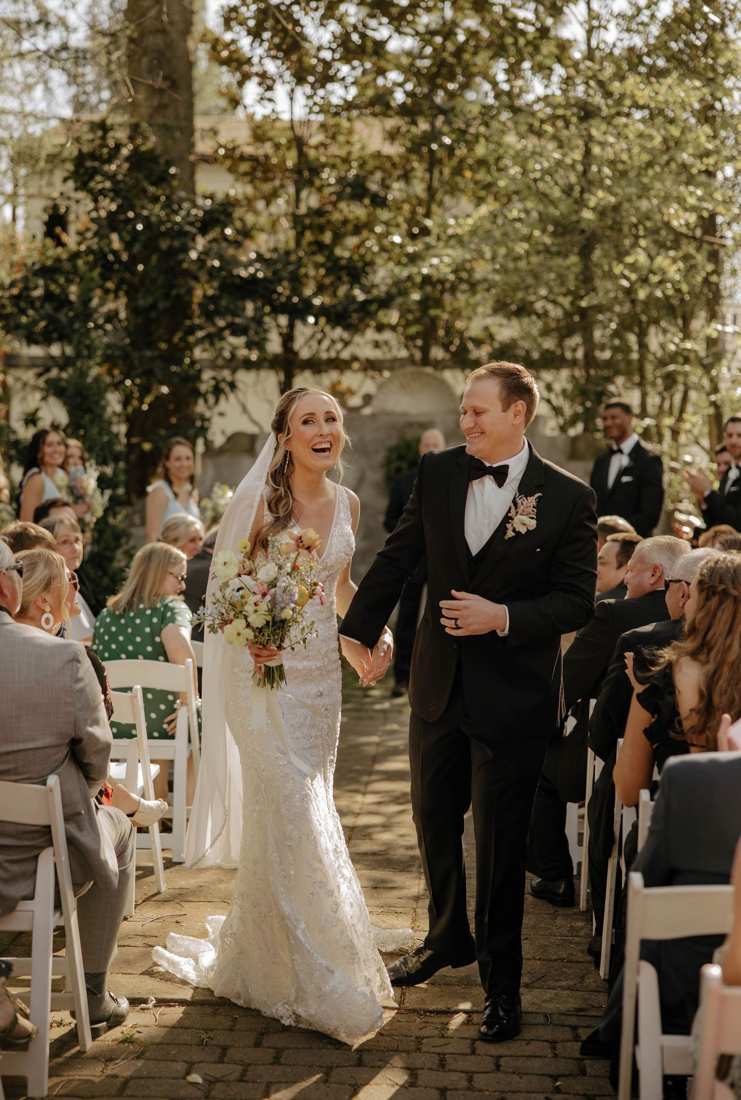A joyful outdoor wedding celebration with guests mingling under trees during a sunny afternoon ceremony.