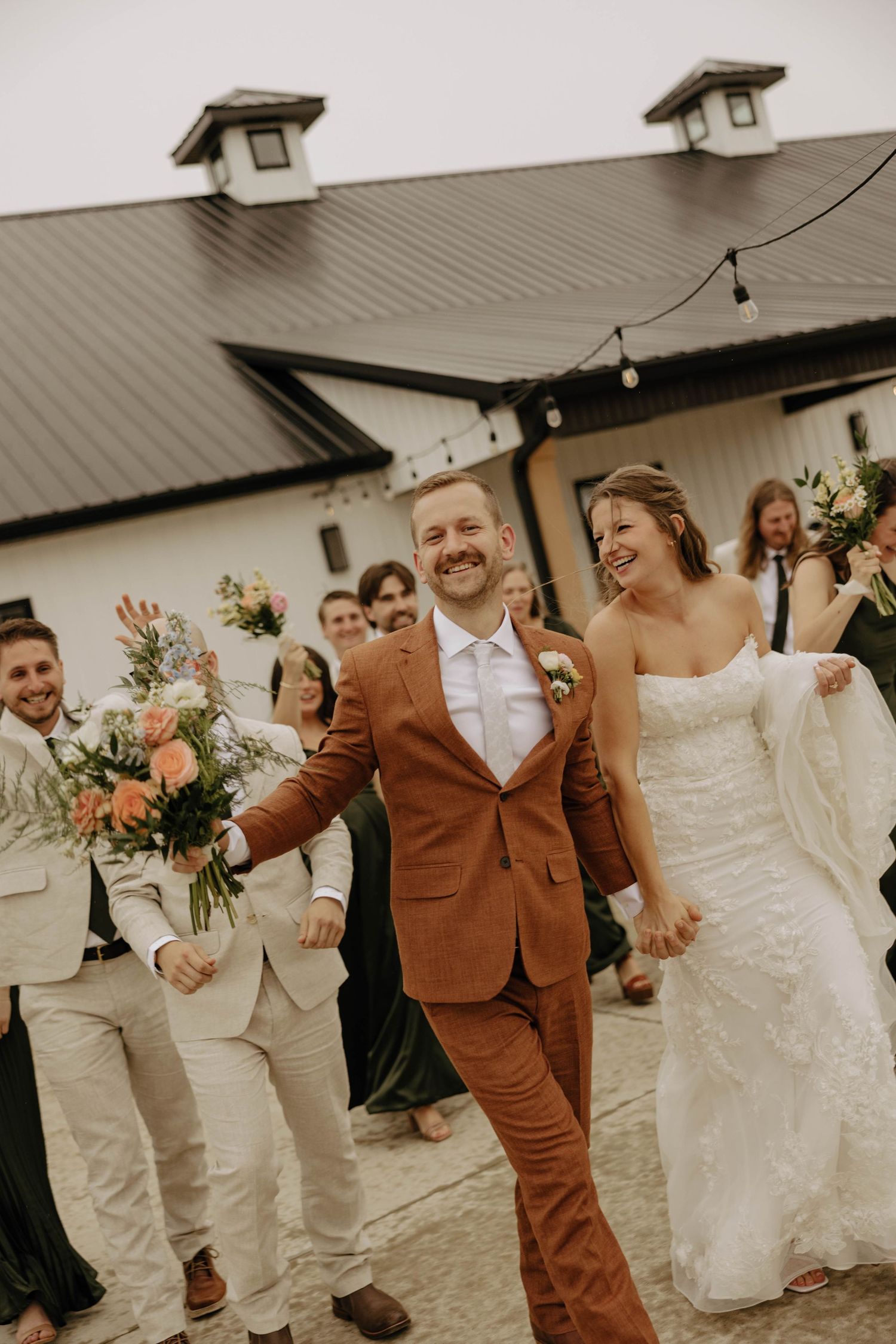 Wedding party in white and brown attire poses together in front of a white building.