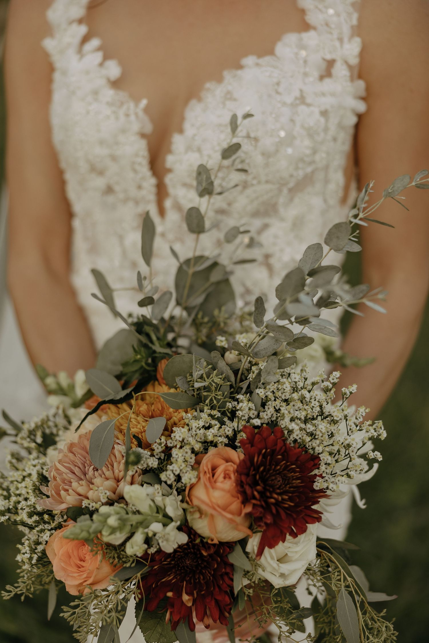 Close-up of a bridal bouquet with burgundy and peach flowers against a lace wedding dress.