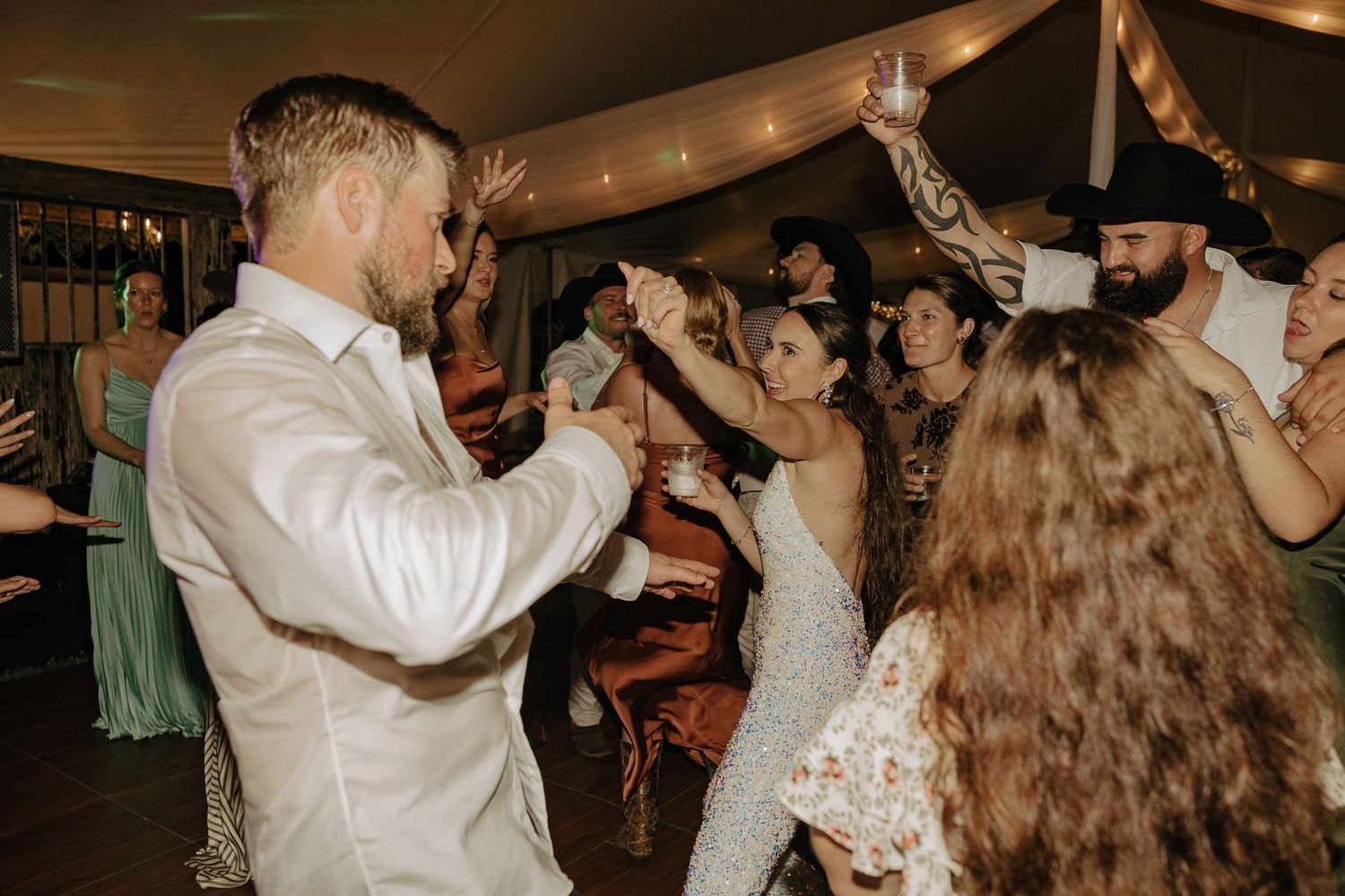 Wedding guests dance enthusiastically under string lights at an indoor reception.