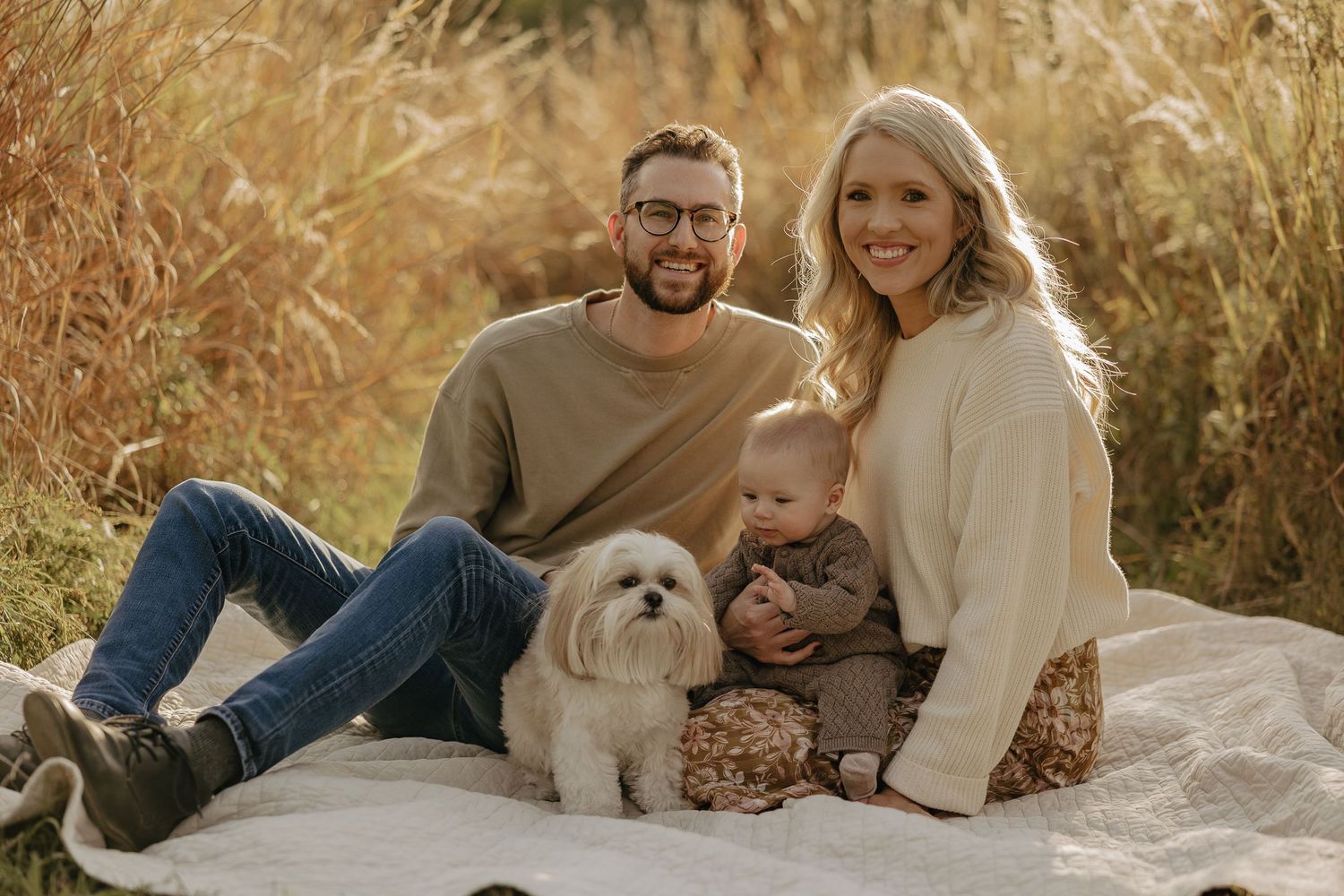 Family photo session with a small white dog on a blanket in tall autumn grass.