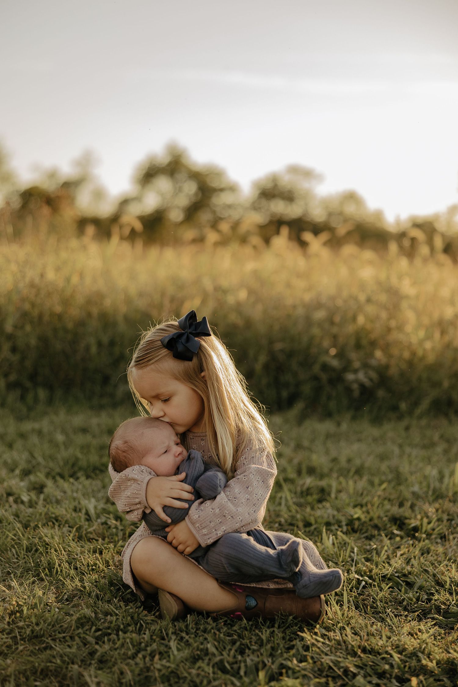 Two children share a tender moment sitting together in a sunlit grassy field at golden hour.