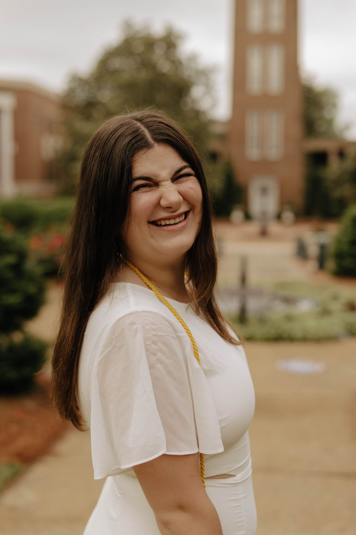 Graduate in white dress poses on campus pathway with brick tower in background.