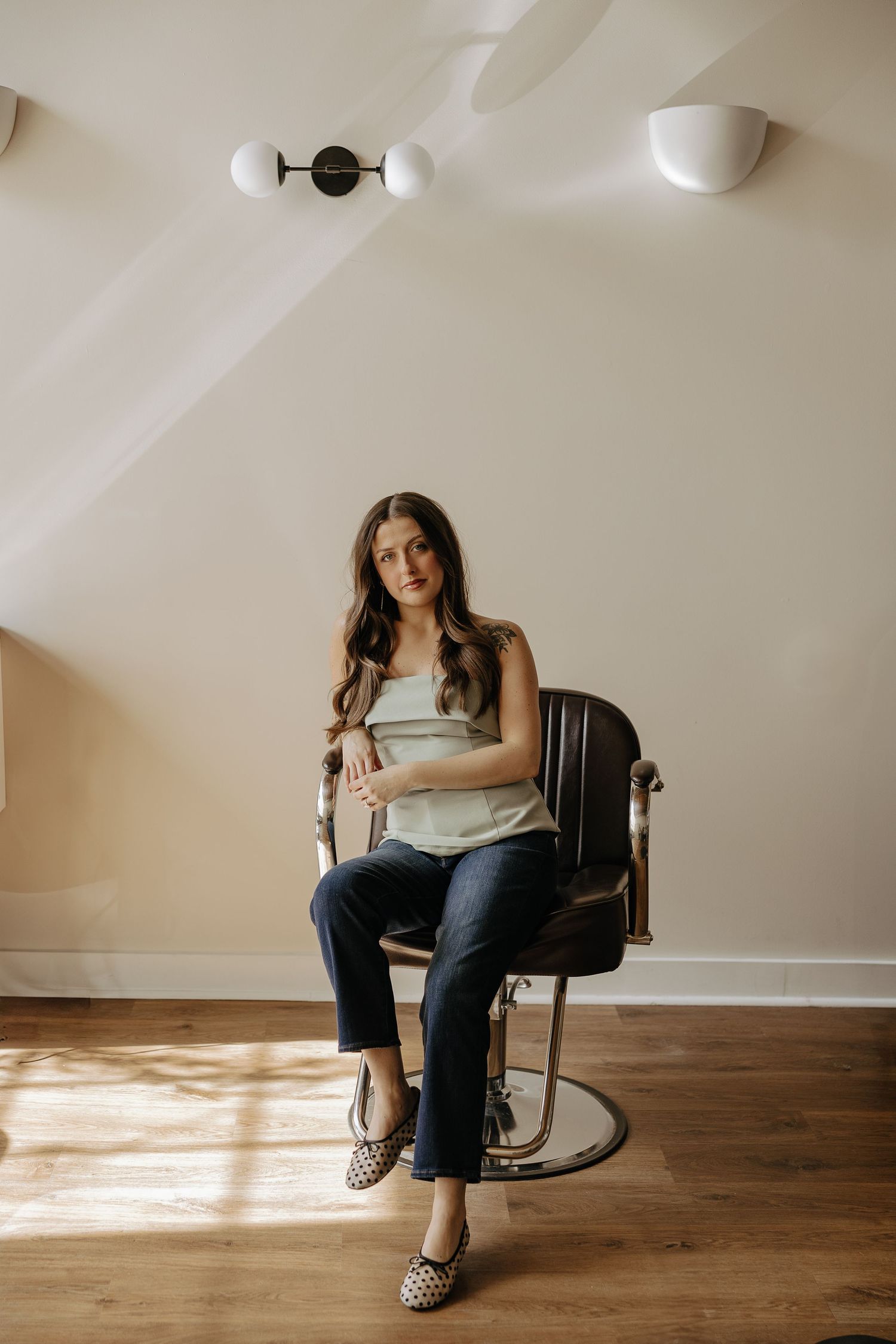 Young woman sitting in a vintage barber chair in a sunlit minimalist room with wood floors.