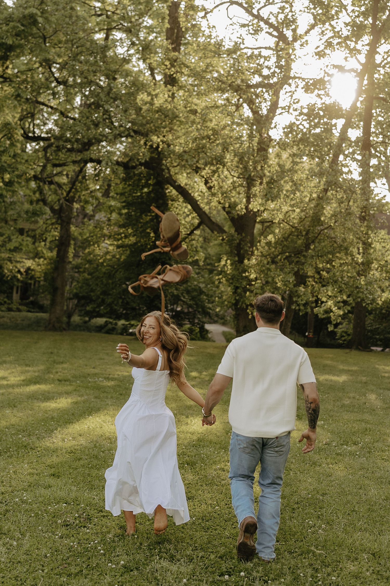 Couple holding hands and running through a lush green park, woman in white dress, man in jeans and white shirt, golden hour.