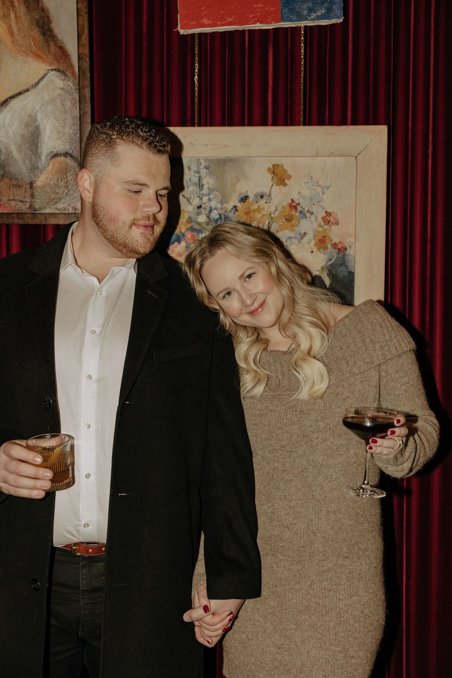 A couple in formal attire share drinks and laughs in front of red curtains.
