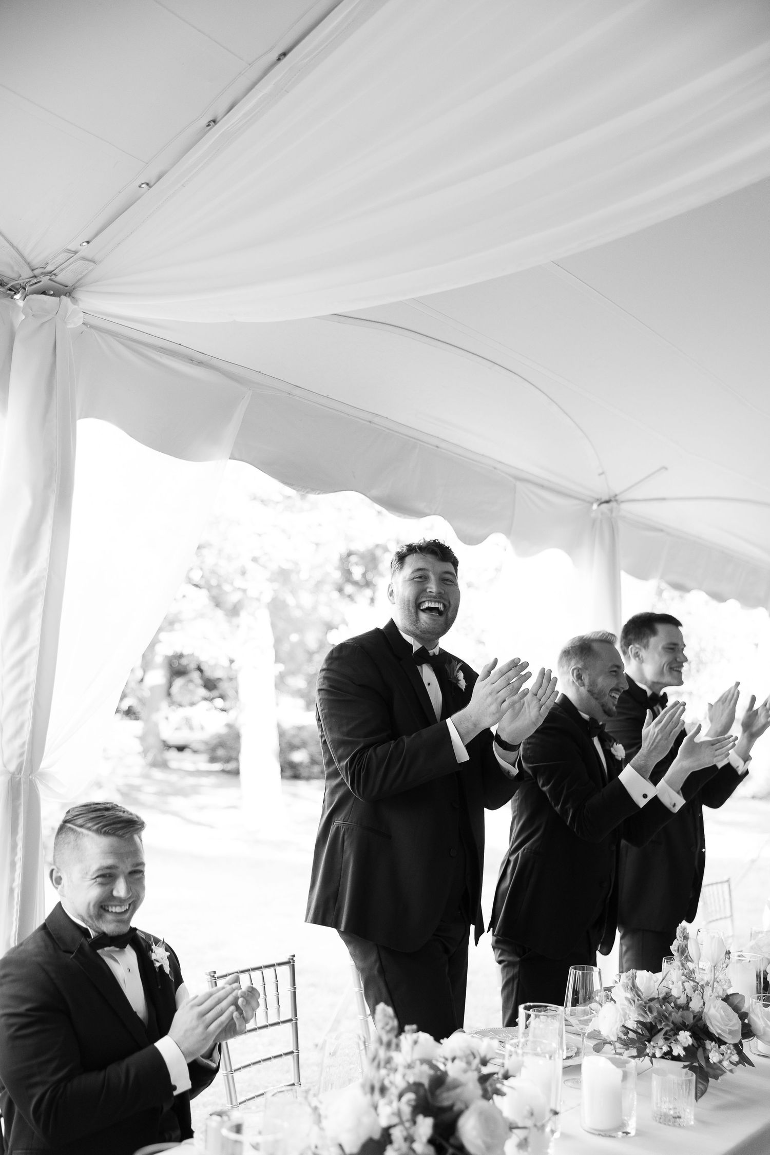 Men in tuxedos clapping and laughing at a wedding reception under a white tent in black and white photo.