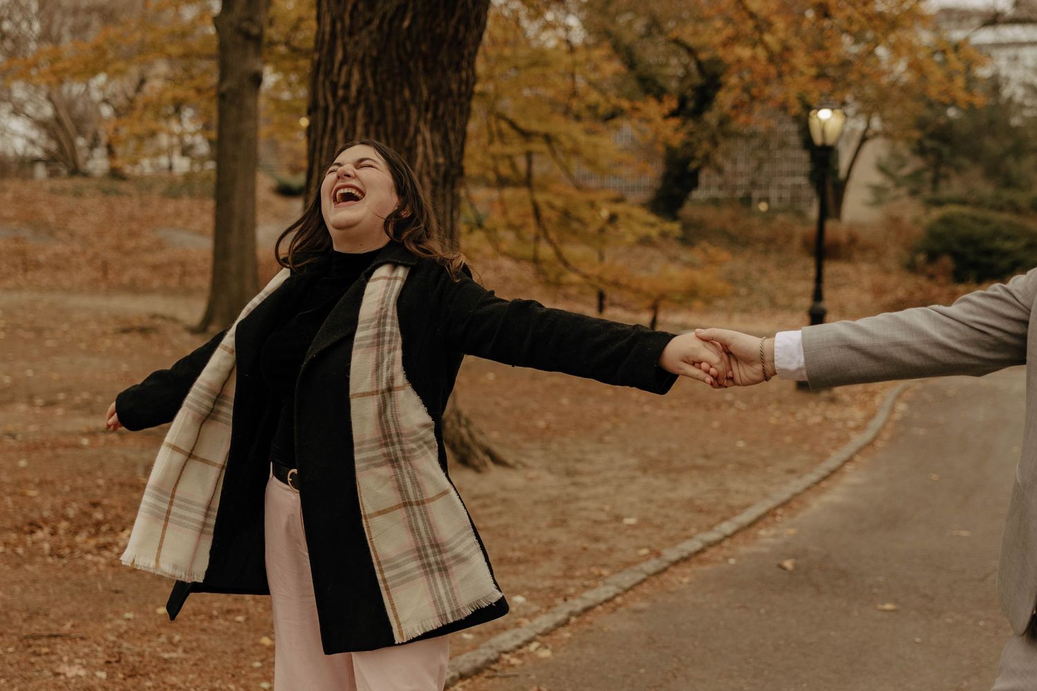 Person in black coat and plaid scarf joyfully spins with outstretched arms on autumn path covered in fallen leaves.