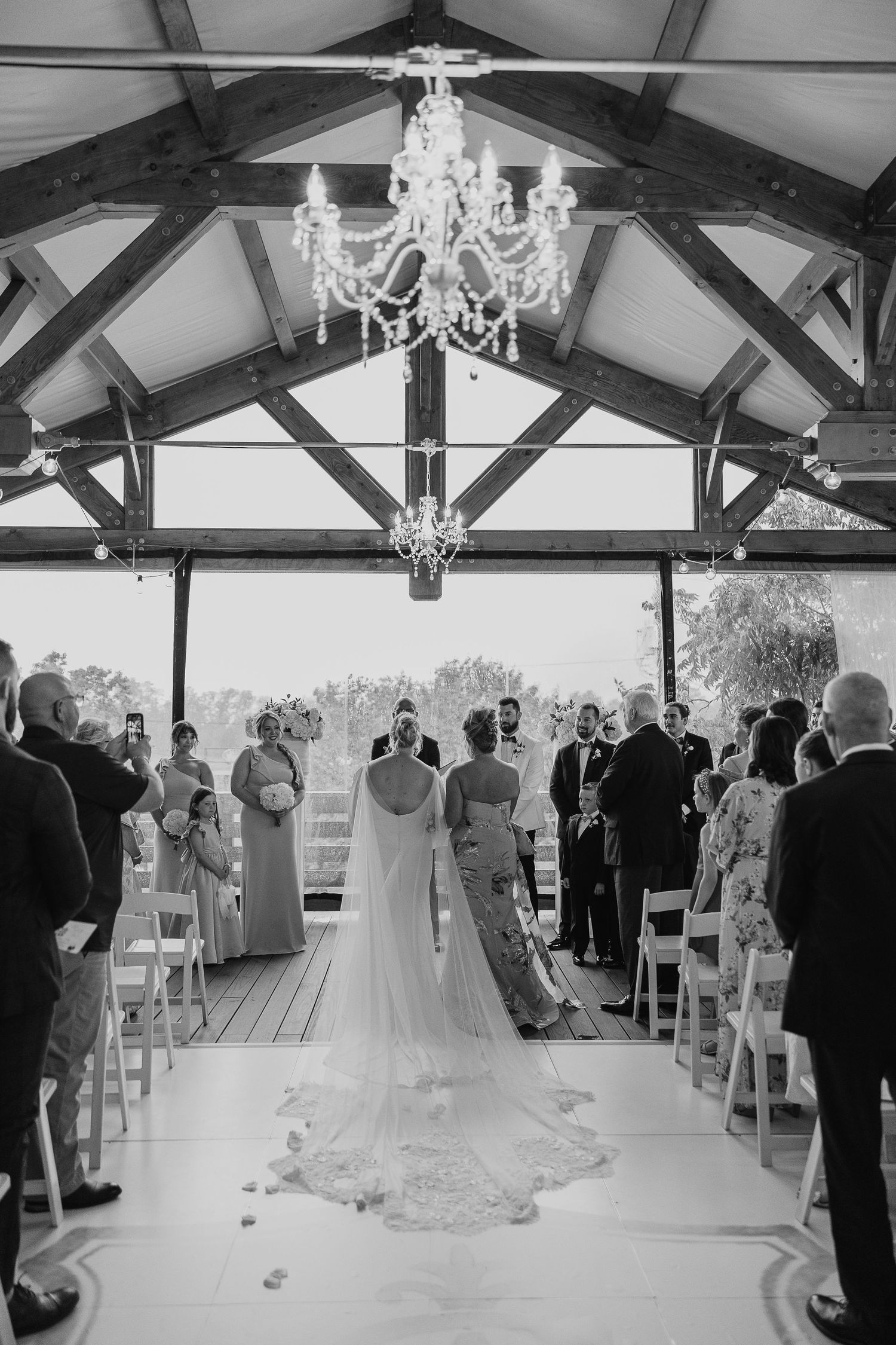 Indoor wedding ceremony setup with exposed beams, crystal chandeliers, and white chairs in a rustic venue.